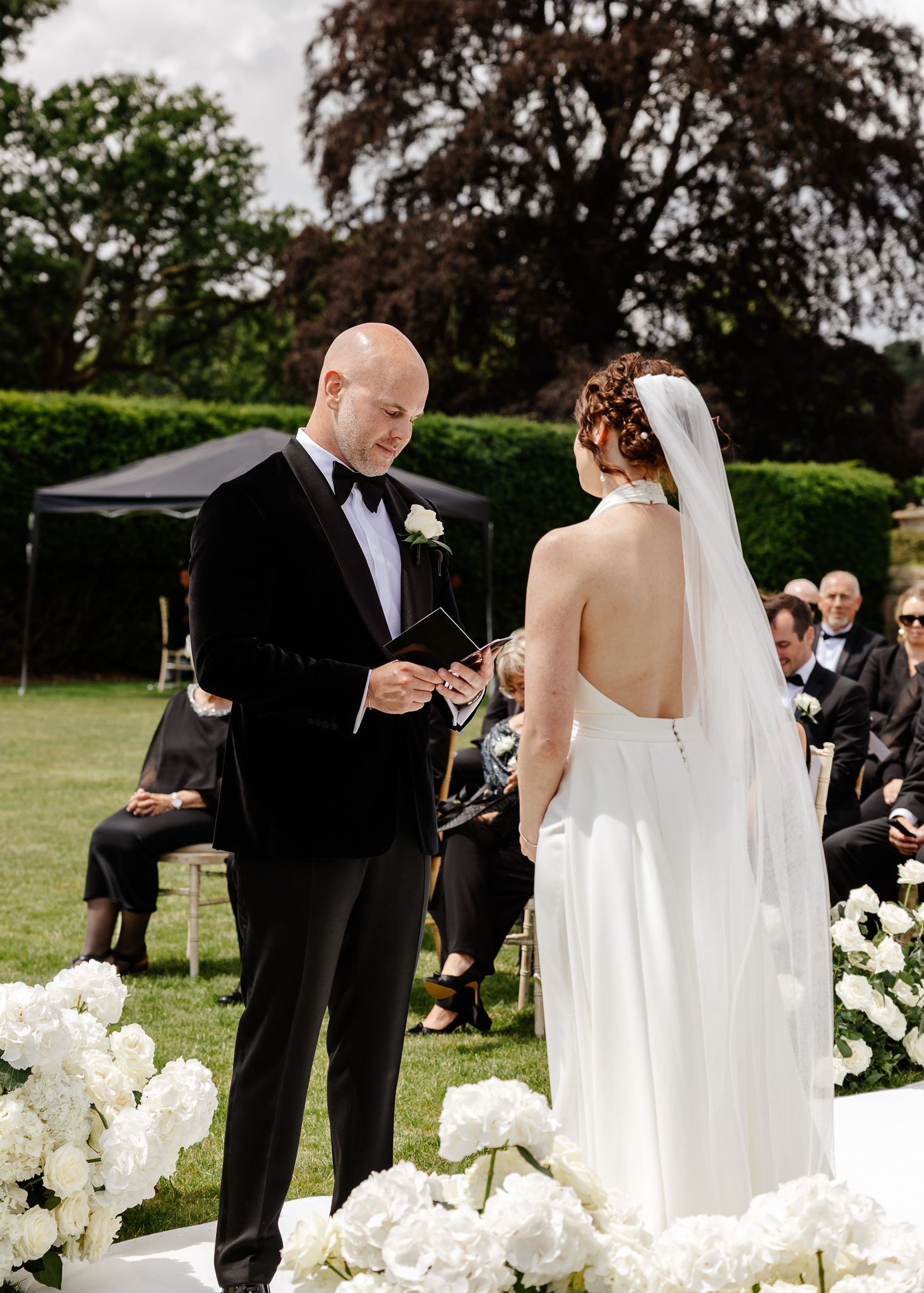 A couple exchanges vows during an outdoor wedding ceremony surrounded by white floral arrangements and seated guests.