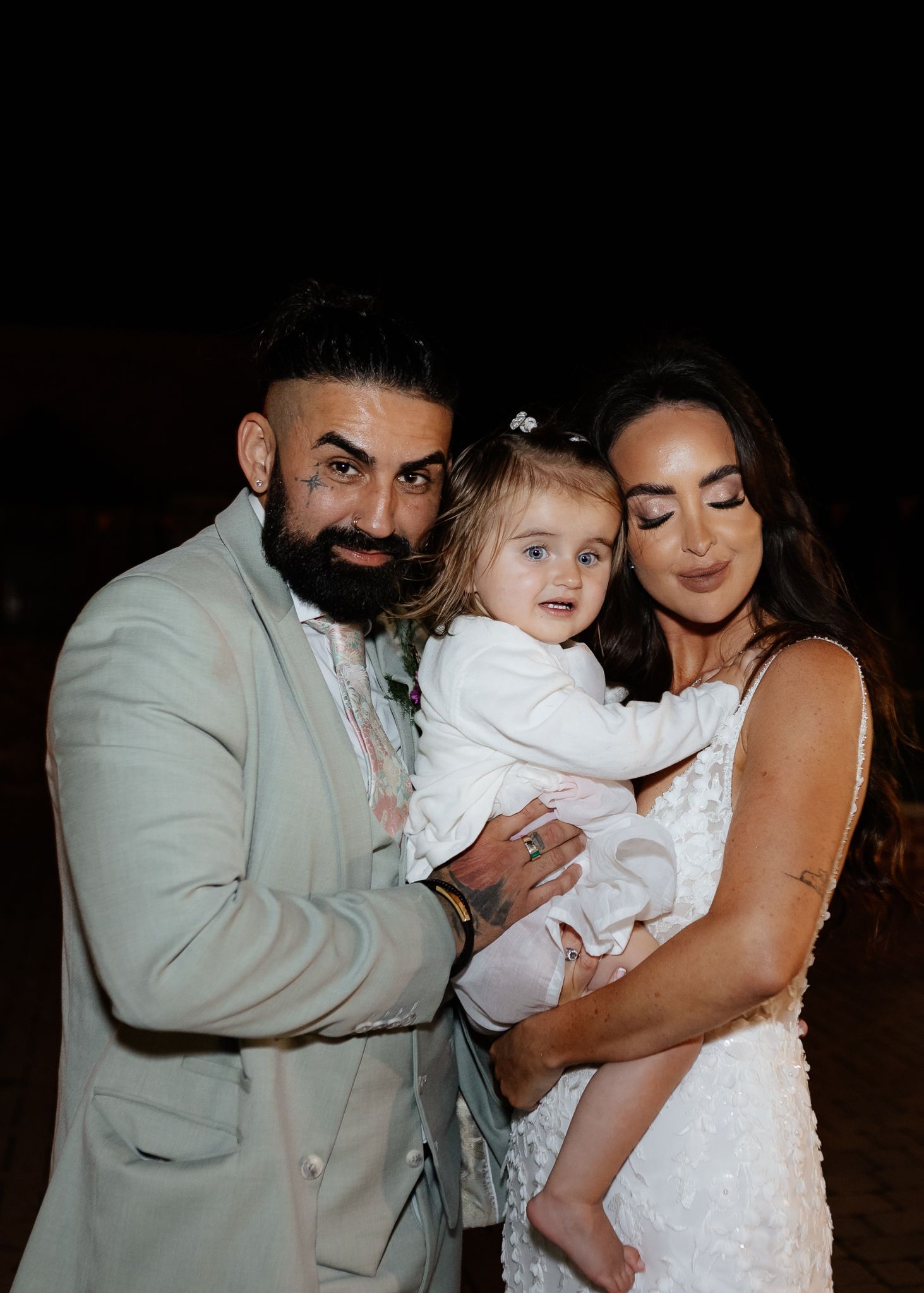 A couple in formal attire embraces a small child in white dress during what appears to be a special occasion.