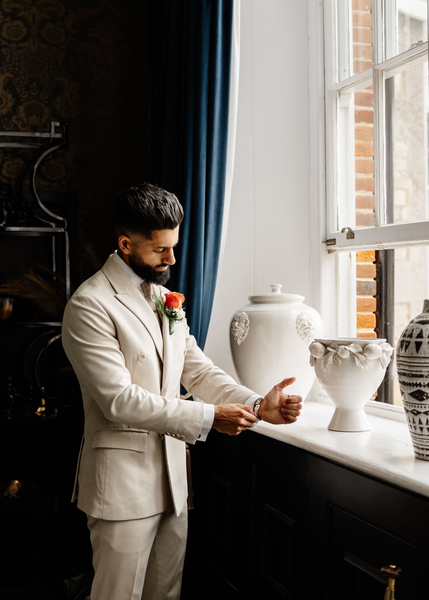 A person in a cream suit reads by a window with decorative vases on the windowsill.
