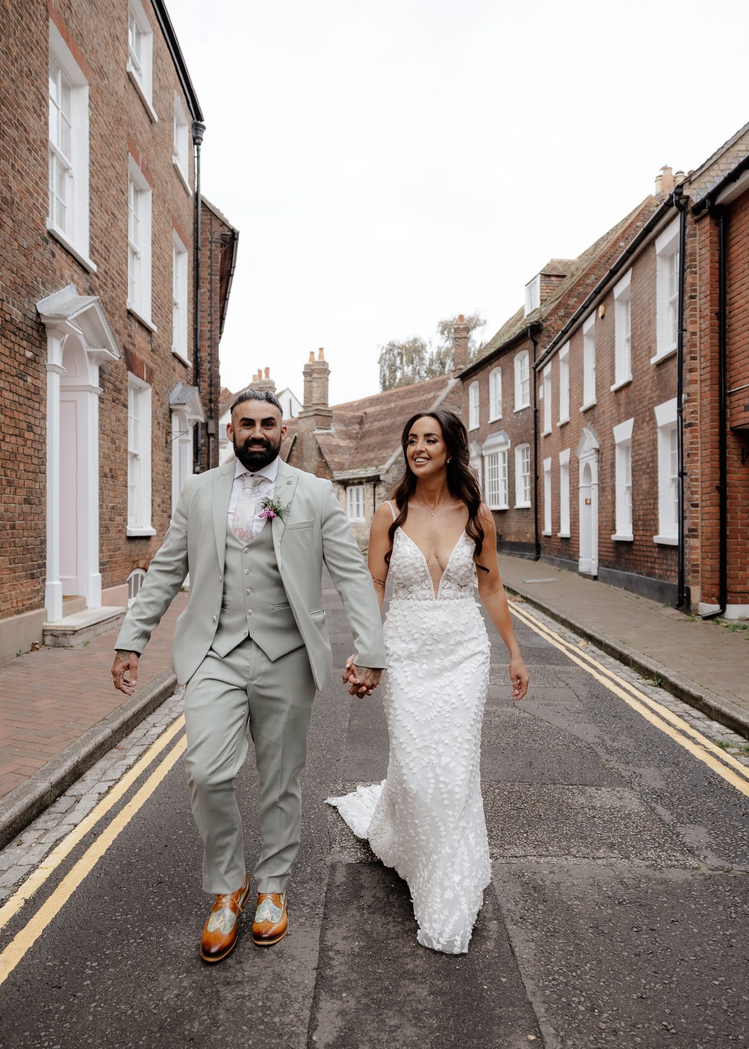 A couple in wedding attire walks down a charming brick-lined street in matching sage green and white outfits.