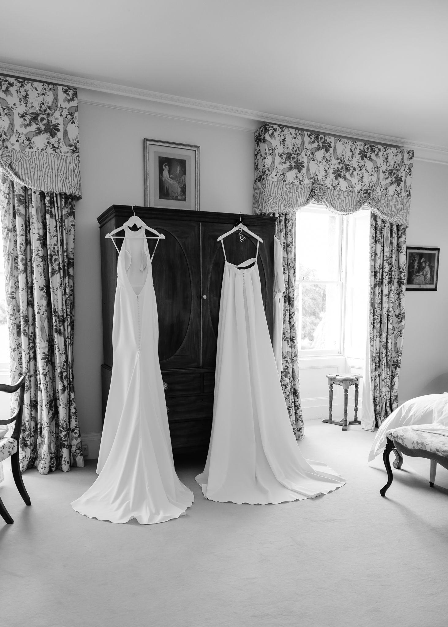 Two elegant white wedding dresses hanging on a dark wardrobe in a room with floral curtains and classic decor.