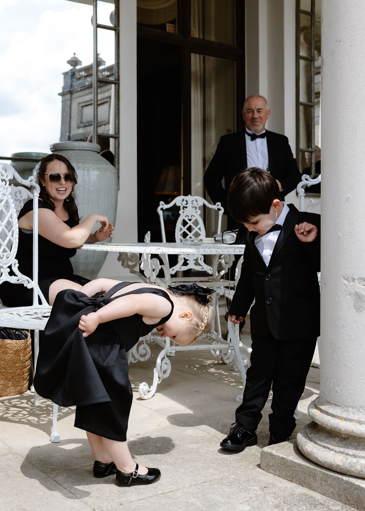 Wedding party members in black attire pose creatively against white columns on a porch.