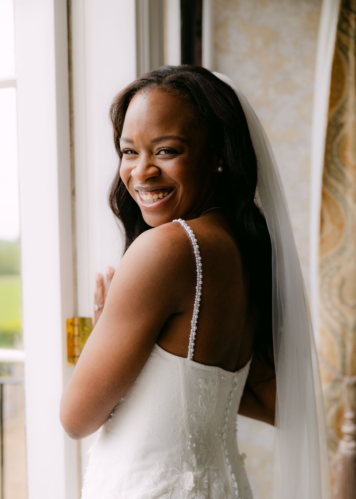 Smiling bride in white spaghetti strap gown and veil poses by a window on her wedding day.
