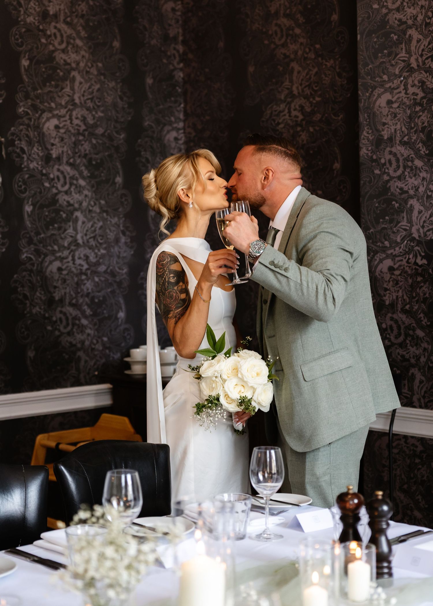 Bride and groom share a kiss while holding champagne glasses at their elegant wedding reception table.