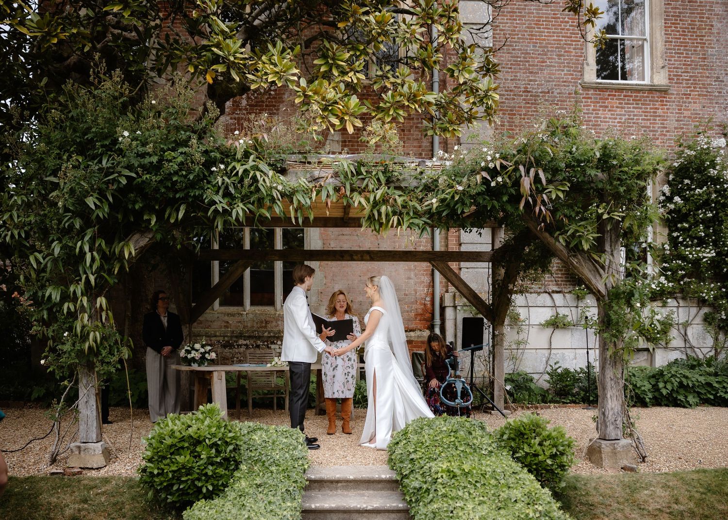 Bride and groom exchange vows outdoors before a charming ivy-covered brick manor house.