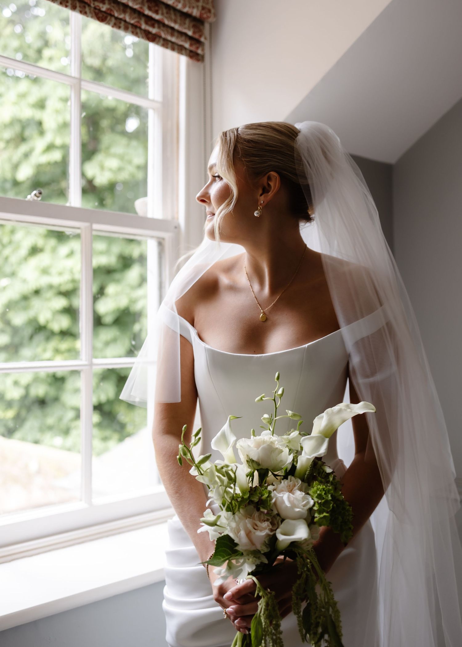 Bride in white gown and veil holds bouquet of white calla lilies, gazing out a sunlit window on her wedding day.