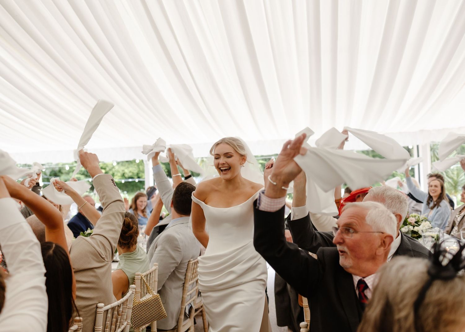 Bride in white gown walks down aisle as guests wave napkins at elegant marquee wedding reception.