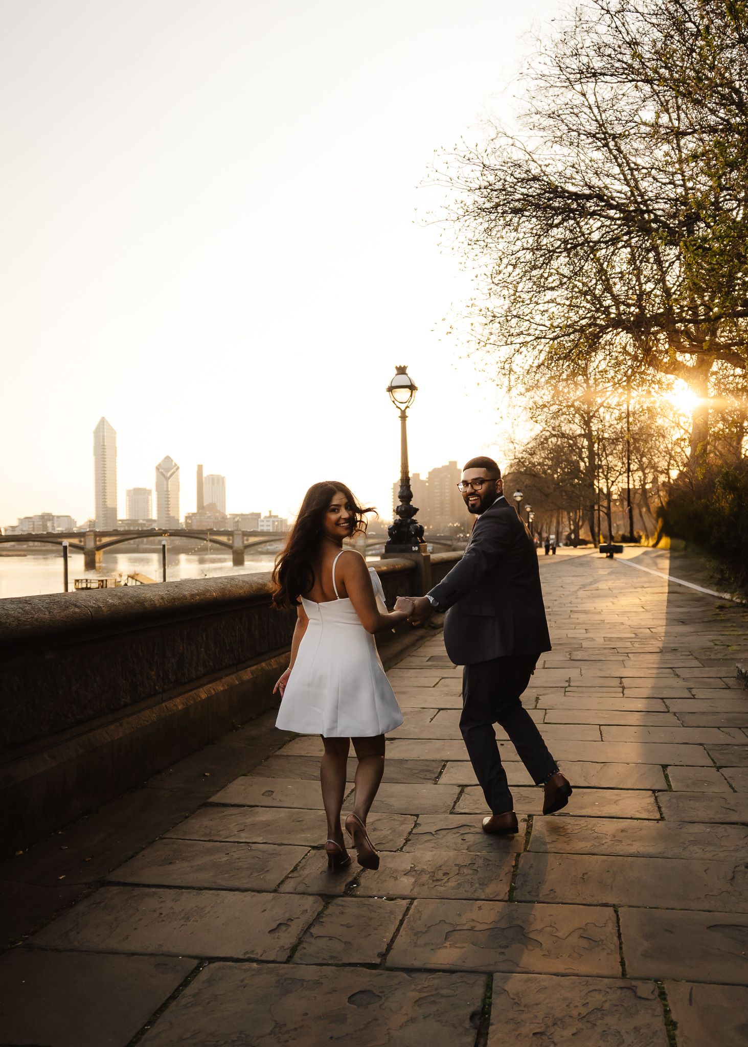 Engaged couple walking along Thames riverbank at golden sunset in London.