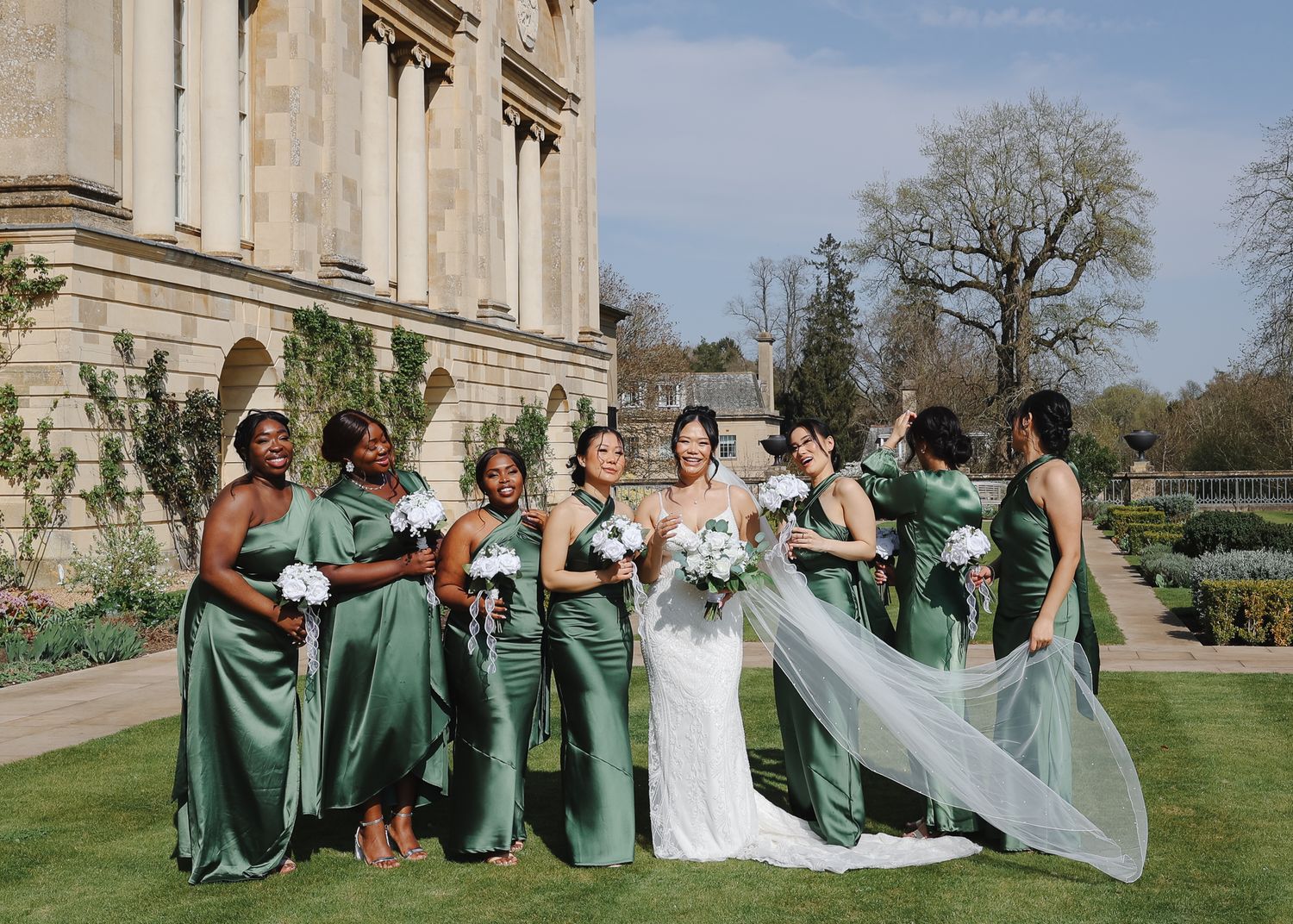 Bridesmaids in emerald green dresses pose with bride's veil on mansion grounds.