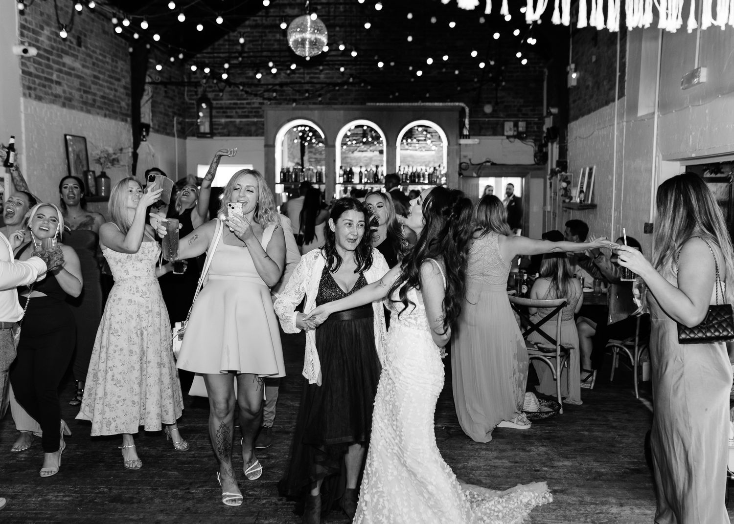 Black and white photo of wedding guests dancing together at a reception in a rustic brick venue.