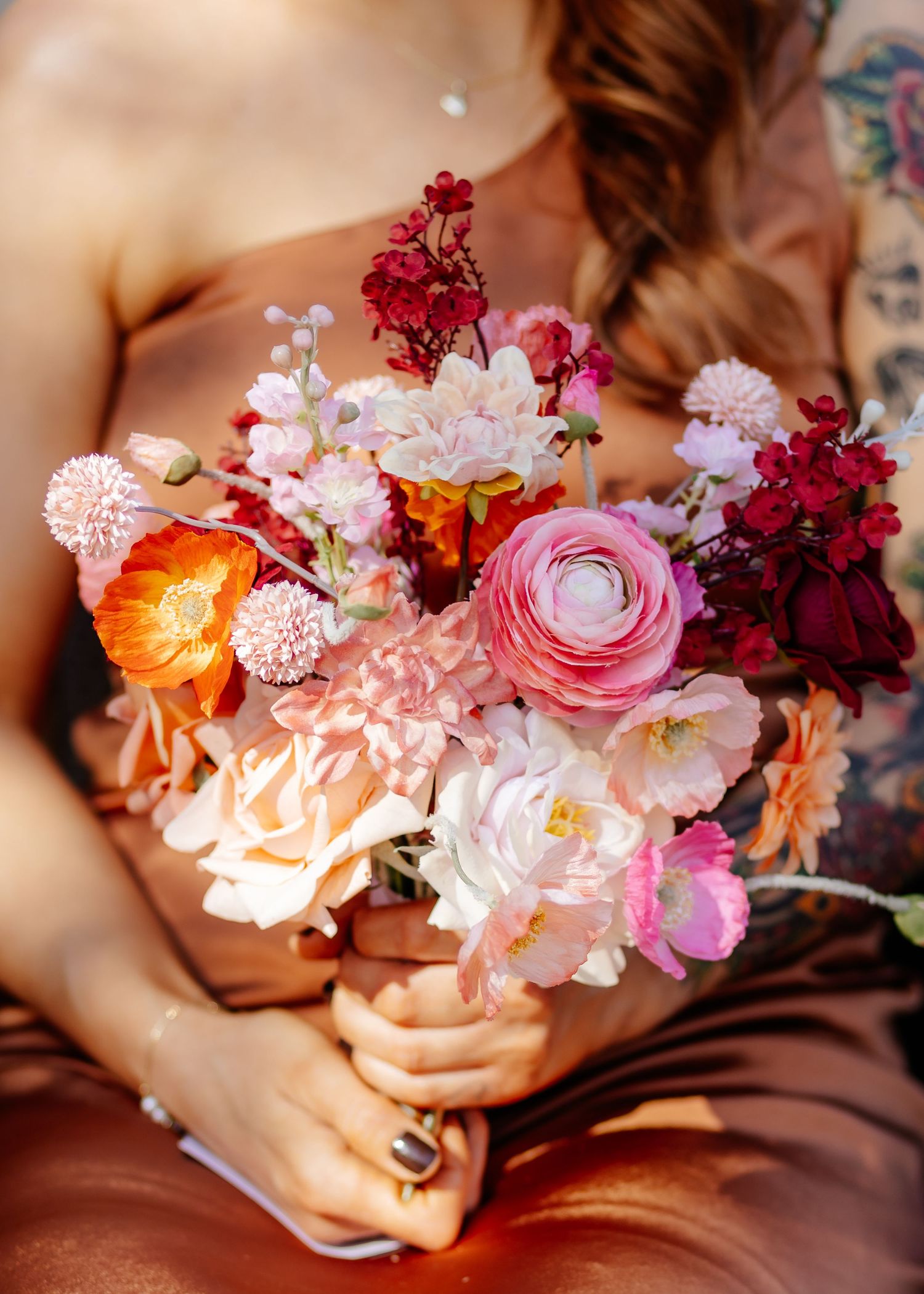 A romantic floral arrangement featuring pink ranunculus, carnations and burgundy blooms held gently in soft lighting.