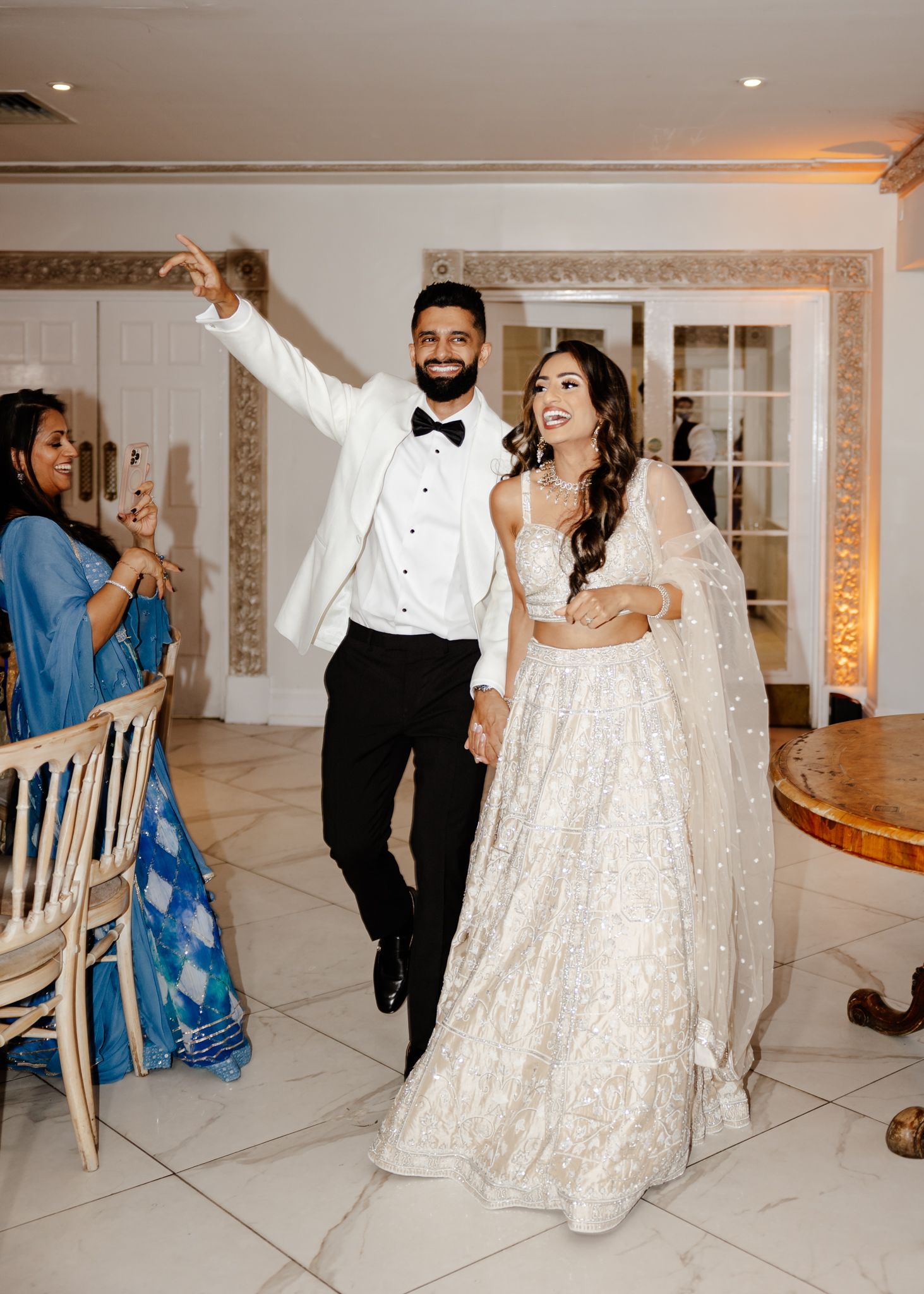 A couple in formal wedding attire dances and celebrates in an elegant white-tiled reception venue at sunset.