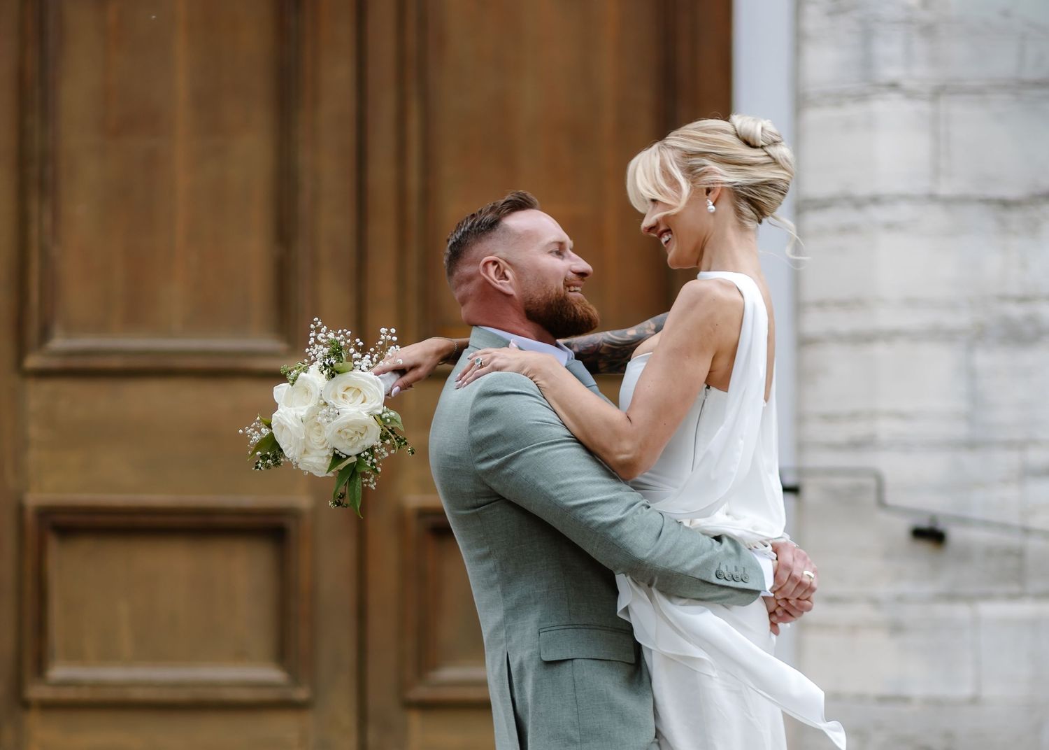 Bride and groom embracing and smiling at each other outdoors, holding a white bouquet, dressed in wedding attire.