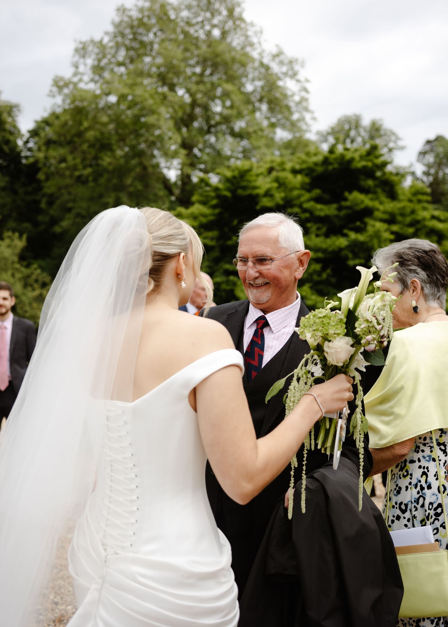 Bride in white gown and veil receives bouquet of white calla lilies from smiling elderly man at outdoor wedding ceremony.