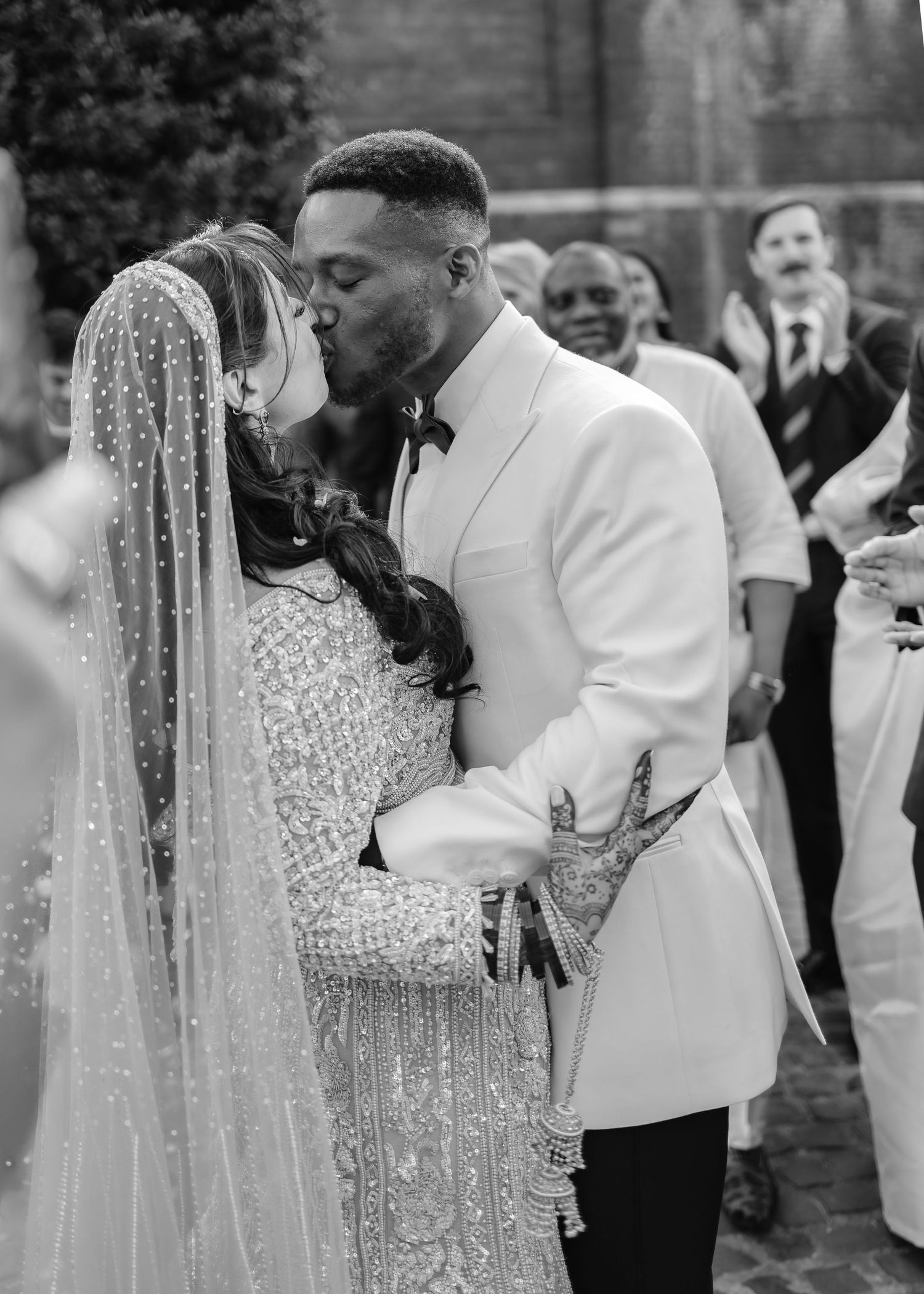 Black and white wedding photo of couple sharing first kiss, bride in beaded gown with veil, groom in white tuxedo jacket.