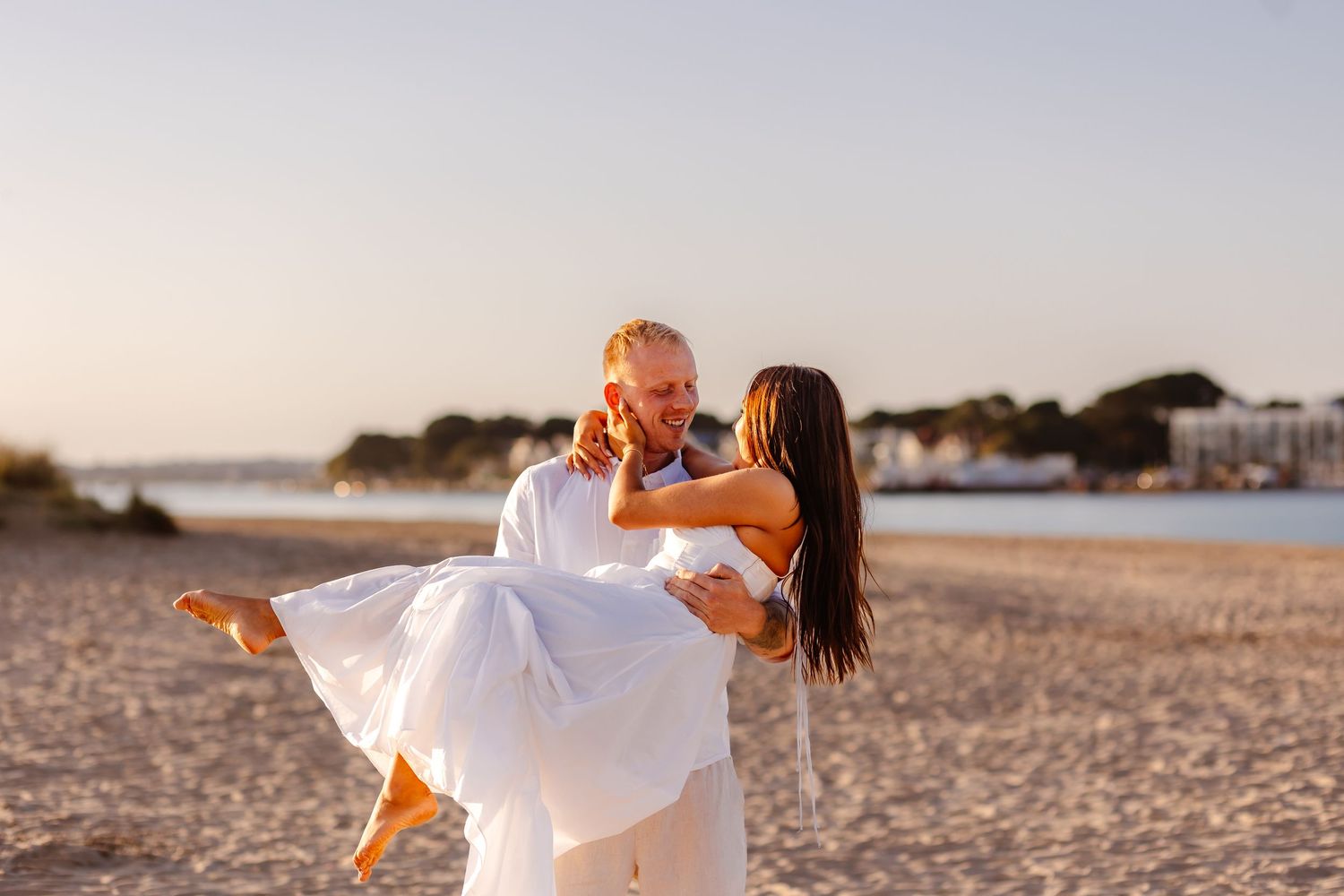 A groom lifts his bride in a white dress on a sunny beach during their wedding celebration.