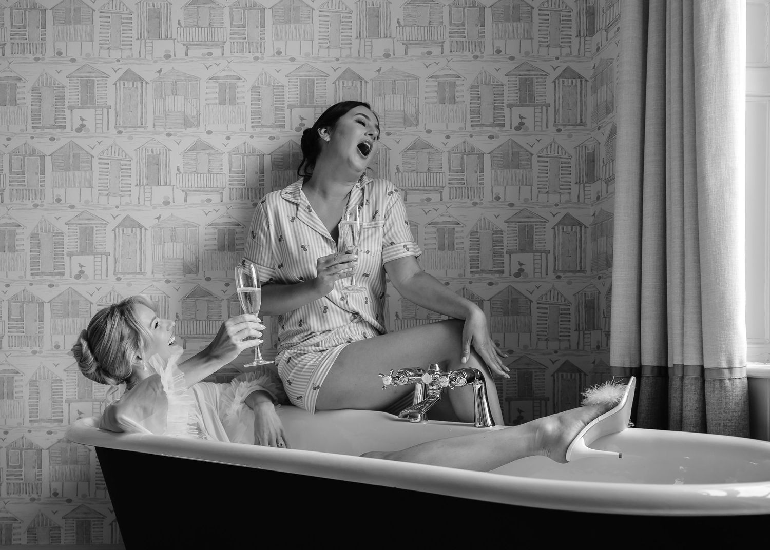 Two women laughing and drinking champagne in a vintage clawfoot bathtub, black and white retro photography.