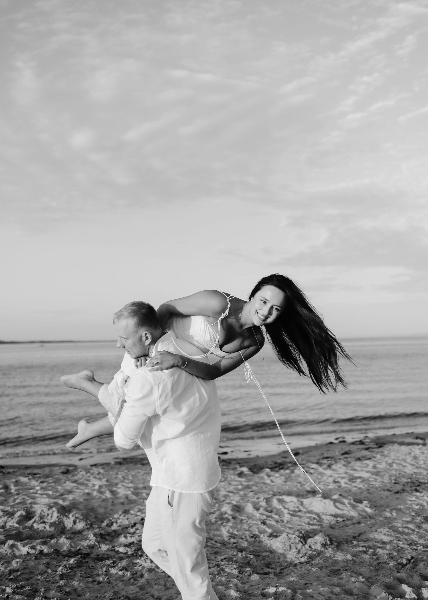 Black and white beach photo of a man carrying a laughing woman on his back, her hair blowing in the wind.