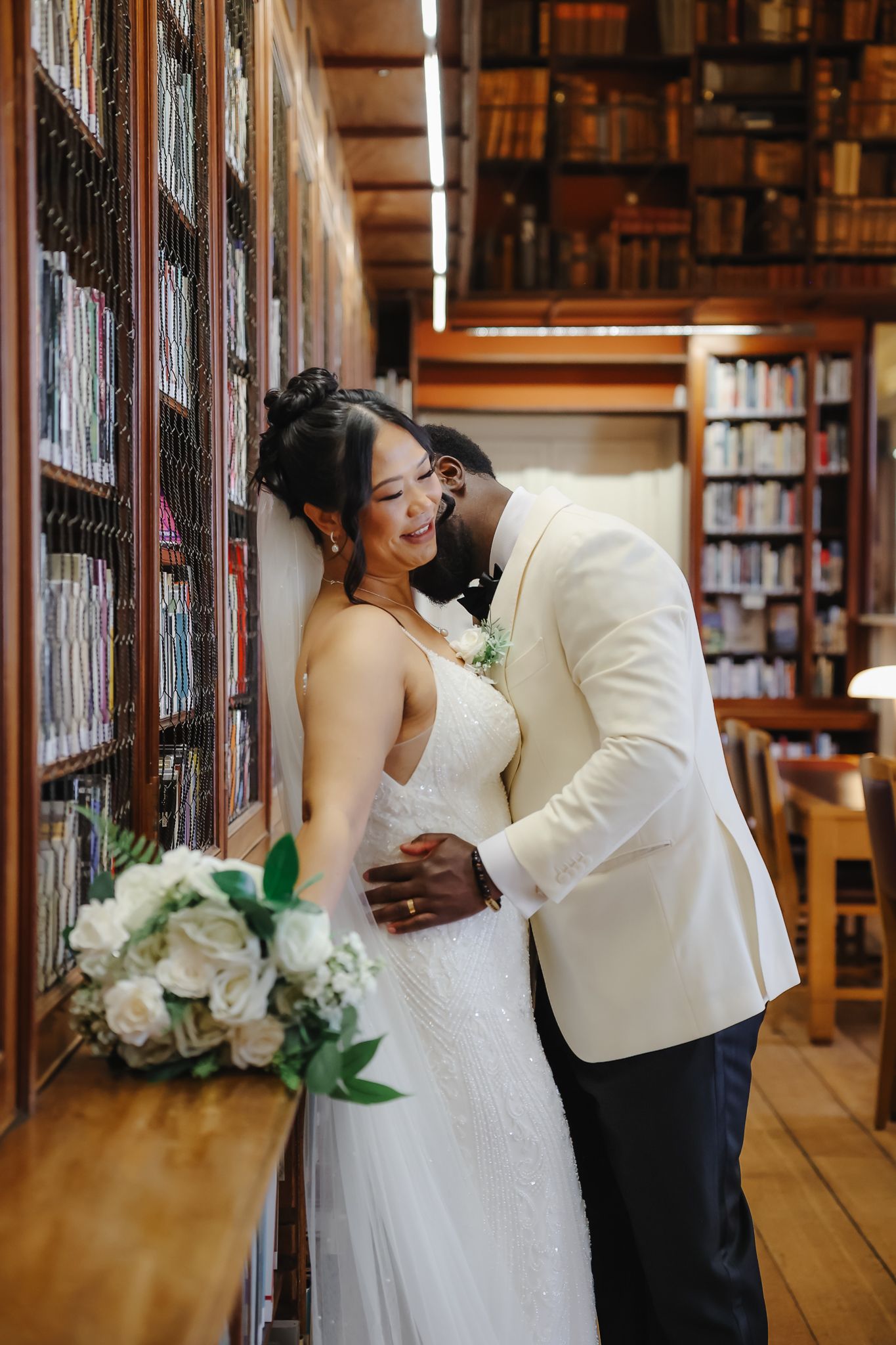 Romantic wedding couple shares intimate moments between library bookshelves with white floral bouquet.