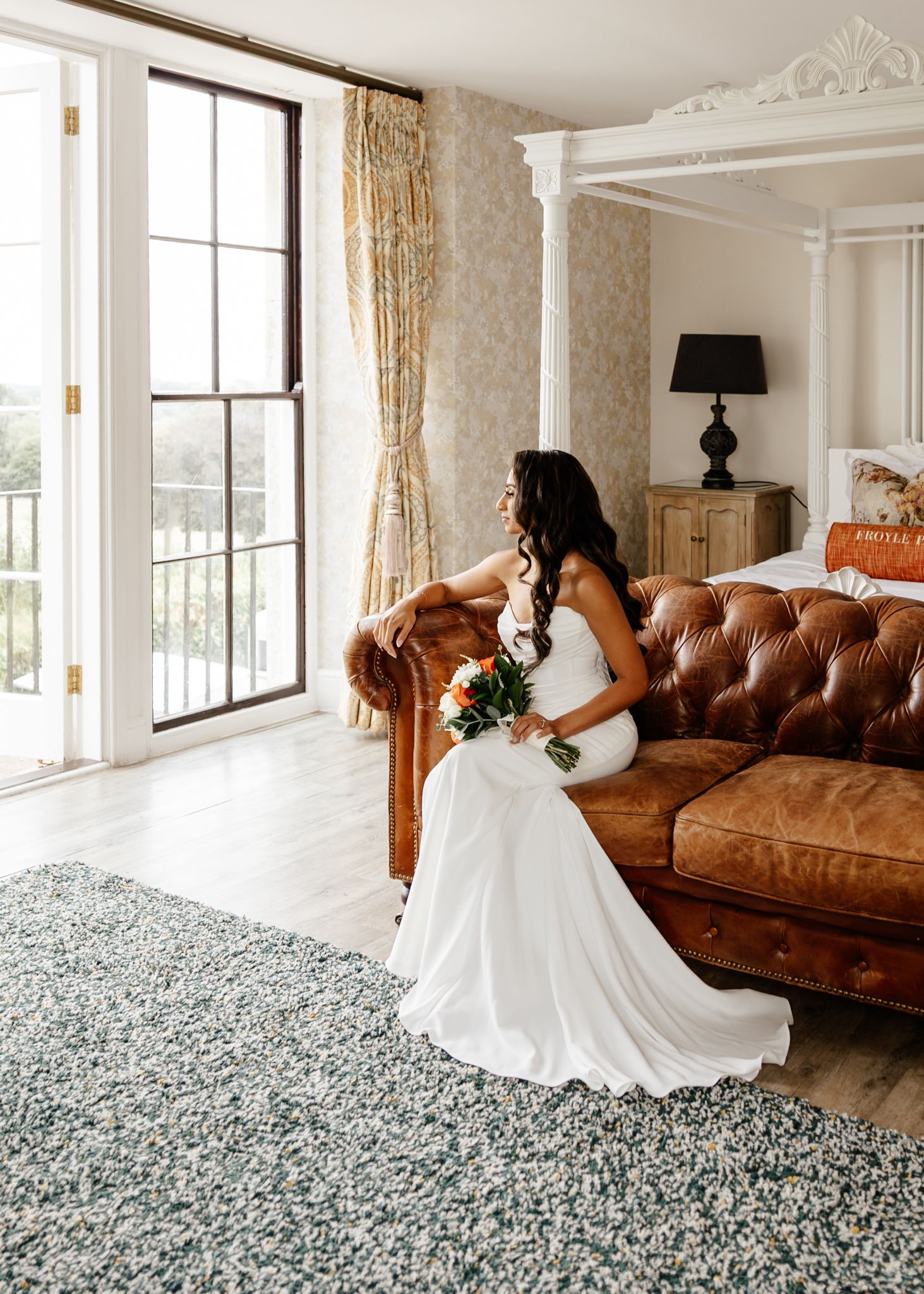 A bride in a flowing white wedding dress poses gracefully on a brown leather couch in an elegant room with large windows.