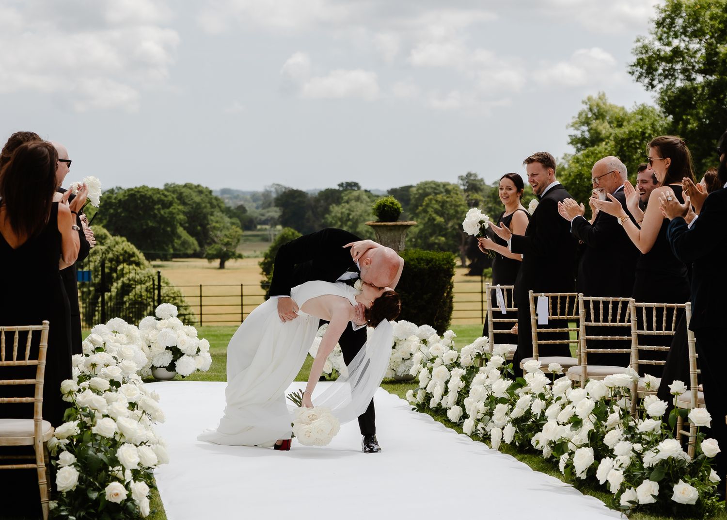 A couple shares a romantic moment on a white aisle runner surrounded by white floral arrangements and guests at an outdoor wedding.