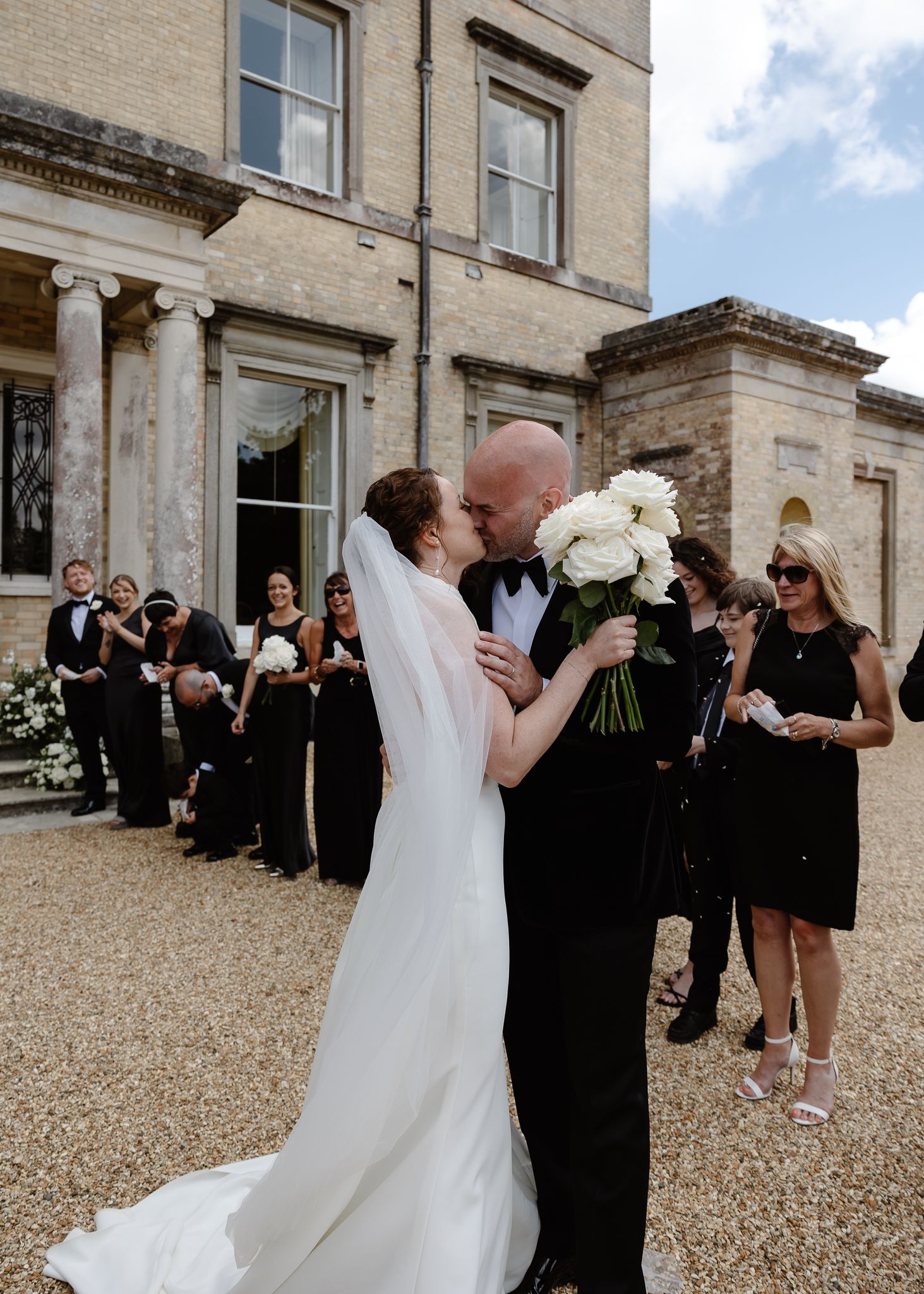 Wedding party gathered outside a grand stone building with classic architecture on a cloudy day.