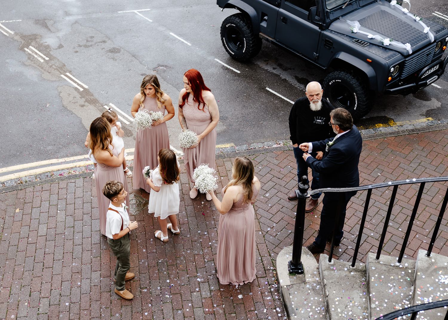 A wedding party group gathers outside on brick pavement near a black car during a celebration.