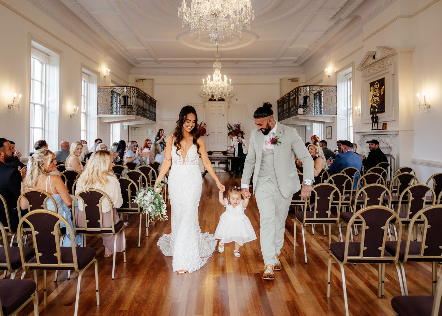 Newlyweds walk down elegant ballroom aisle with flower girl in white dress under crystal chandeliers.