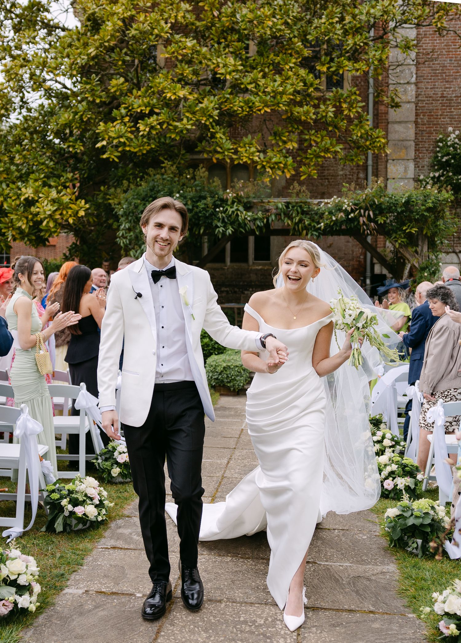 Bride and groom walk joyfully down outdoor aisle, she in white gown with veil, he in white tuxedo jacket.