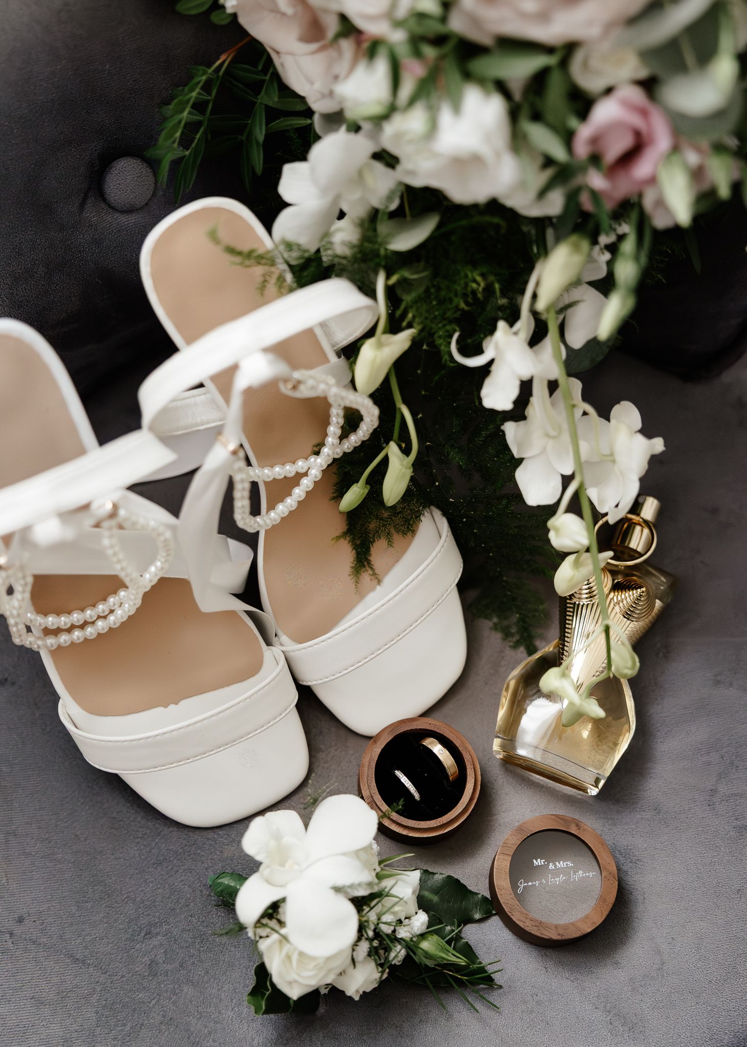 Wedding details featuring white platform heels, delicate pearl jewelry, and gold wedding rings arranged with white floral decor.
