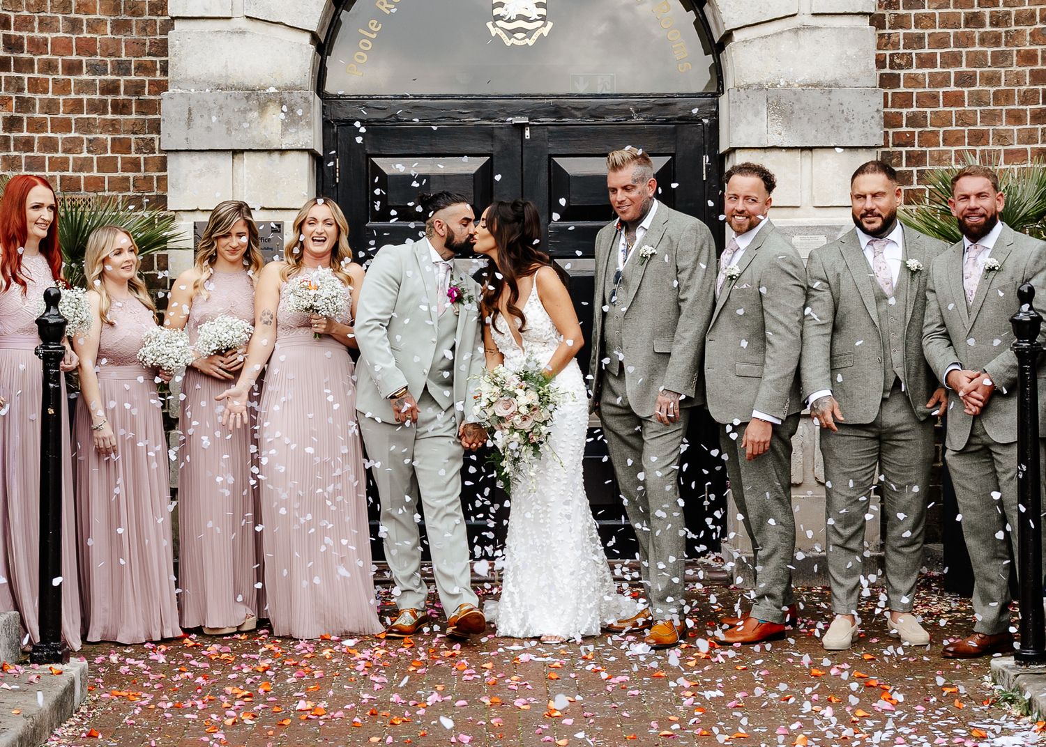 Wedding party in gray suits and pink dresses celebrates with confetti in front of a brick church entrance.