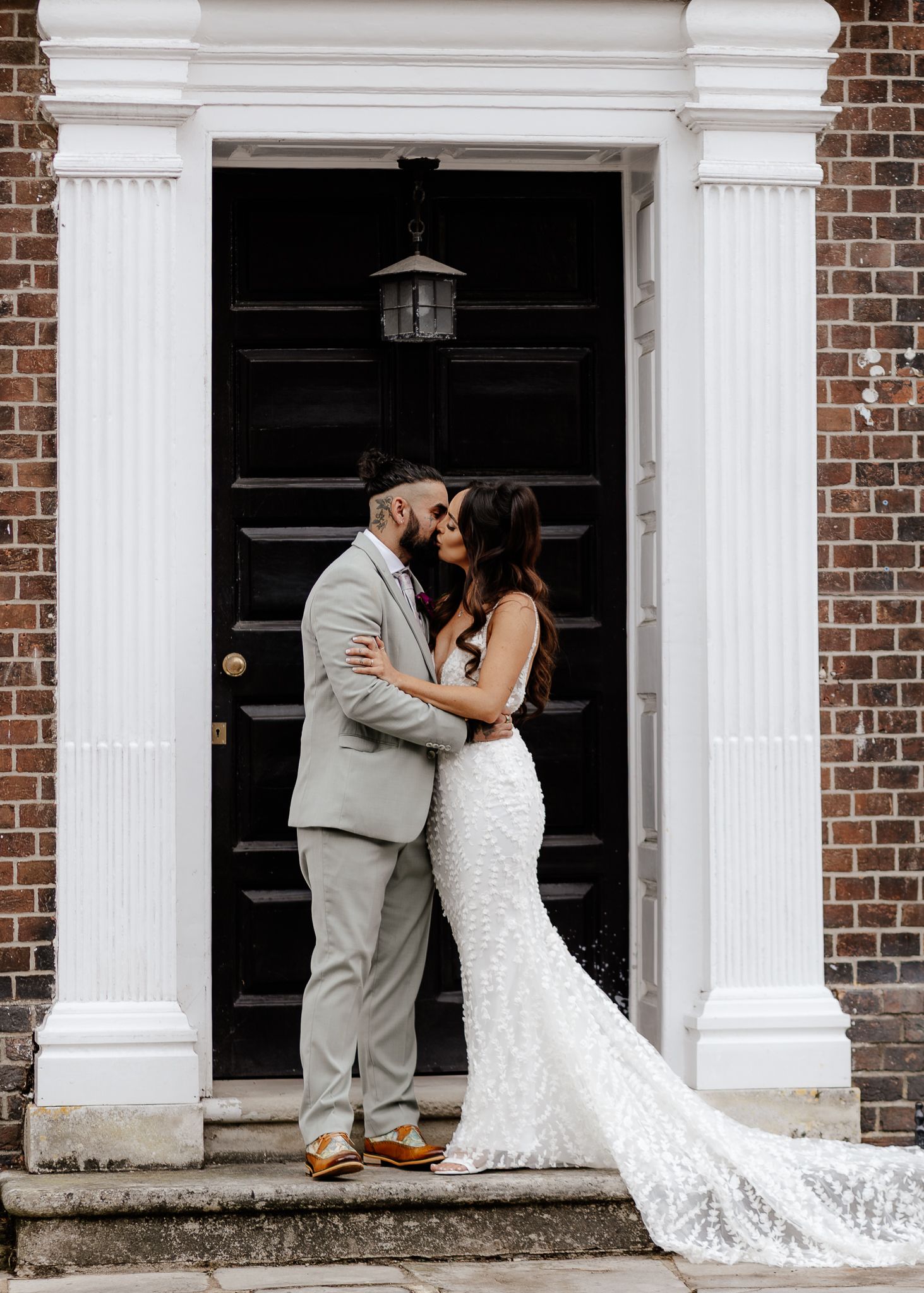 A couple shares a romantic kiss on the steps of a brick building with white columns, the bride's lace dress trailing behind.
