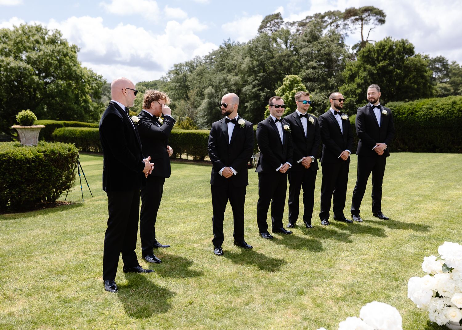 A panoramic sequence of wedding party members in black suits standing in a lush green garden setting.