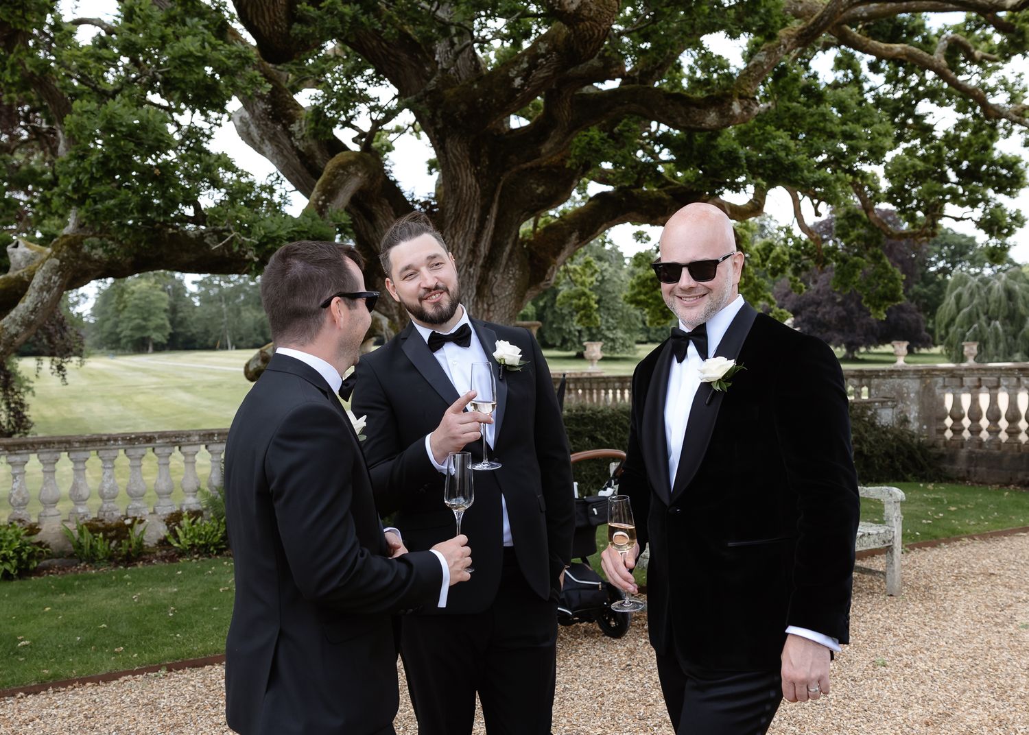 Three men in tuxedos share drinks and laughter in a garden setting.