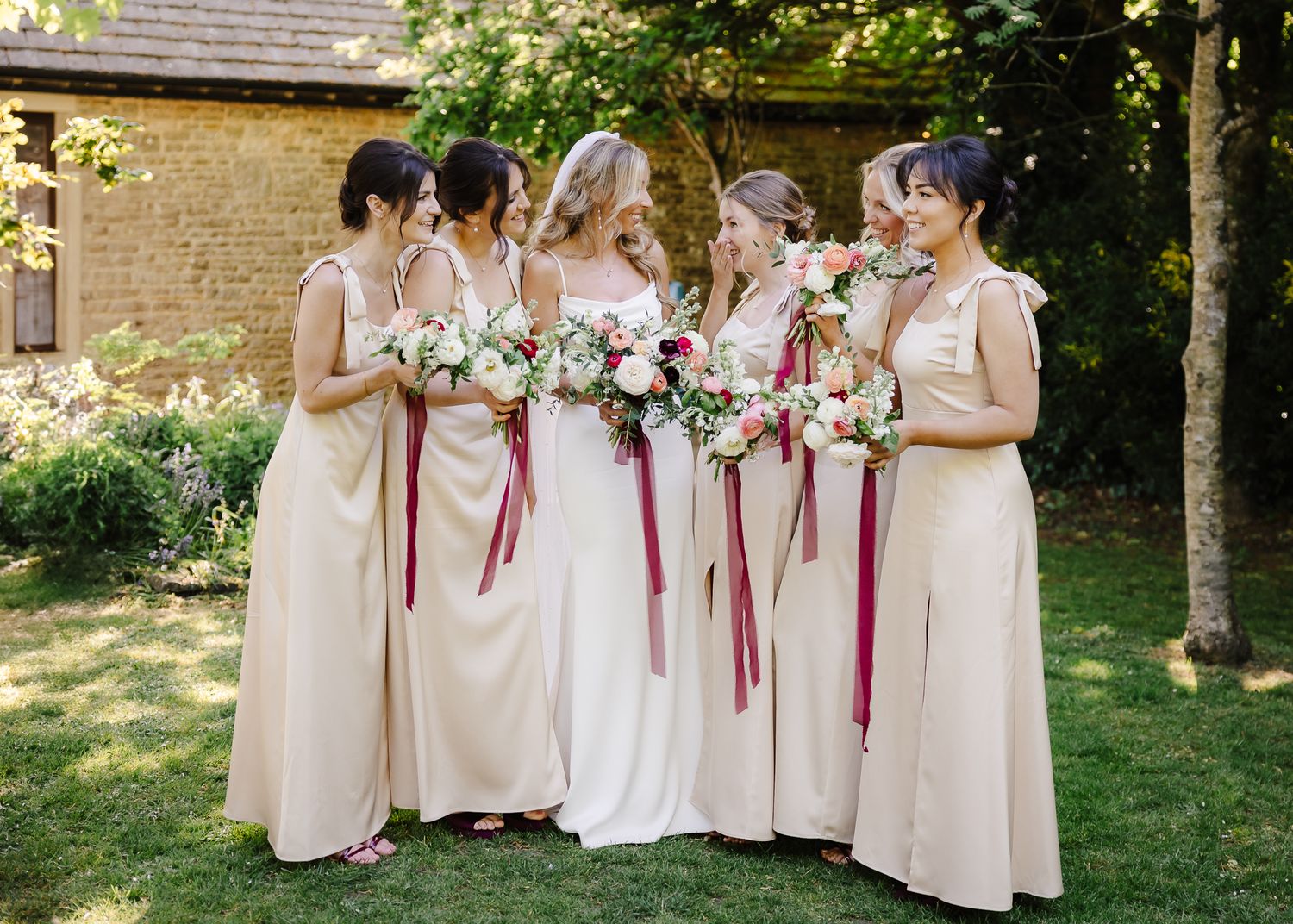 Bridesmaids in blush pink dresses stand together holding burgundy and pink bouquets in an outdoor garden setting.
