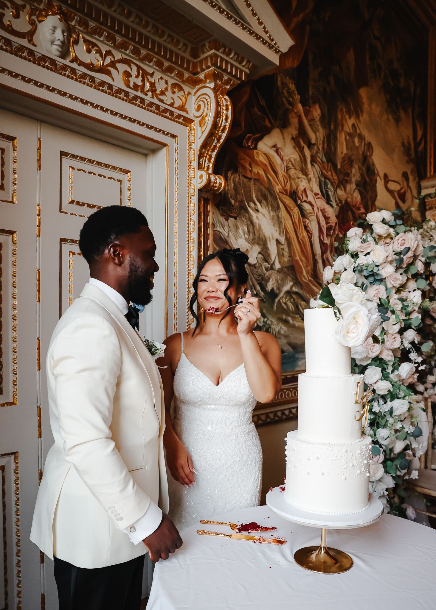Wedding celebration in an ornate room with white decor, floral arrangements, and a tiered white cake.