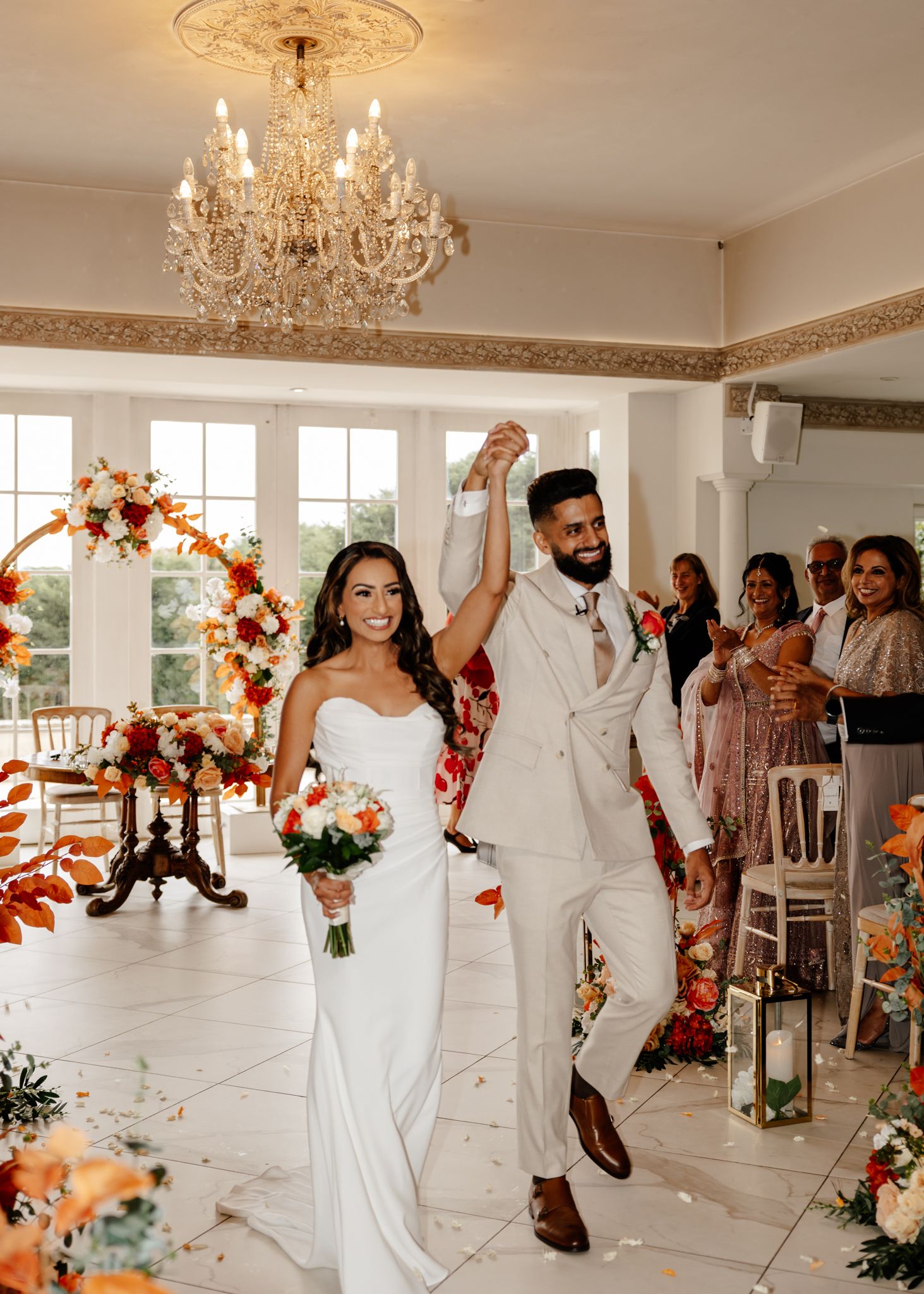 Wedding party in white attire dances and celebrates on an elegant ballroom floor beneath crystal chandeliers.