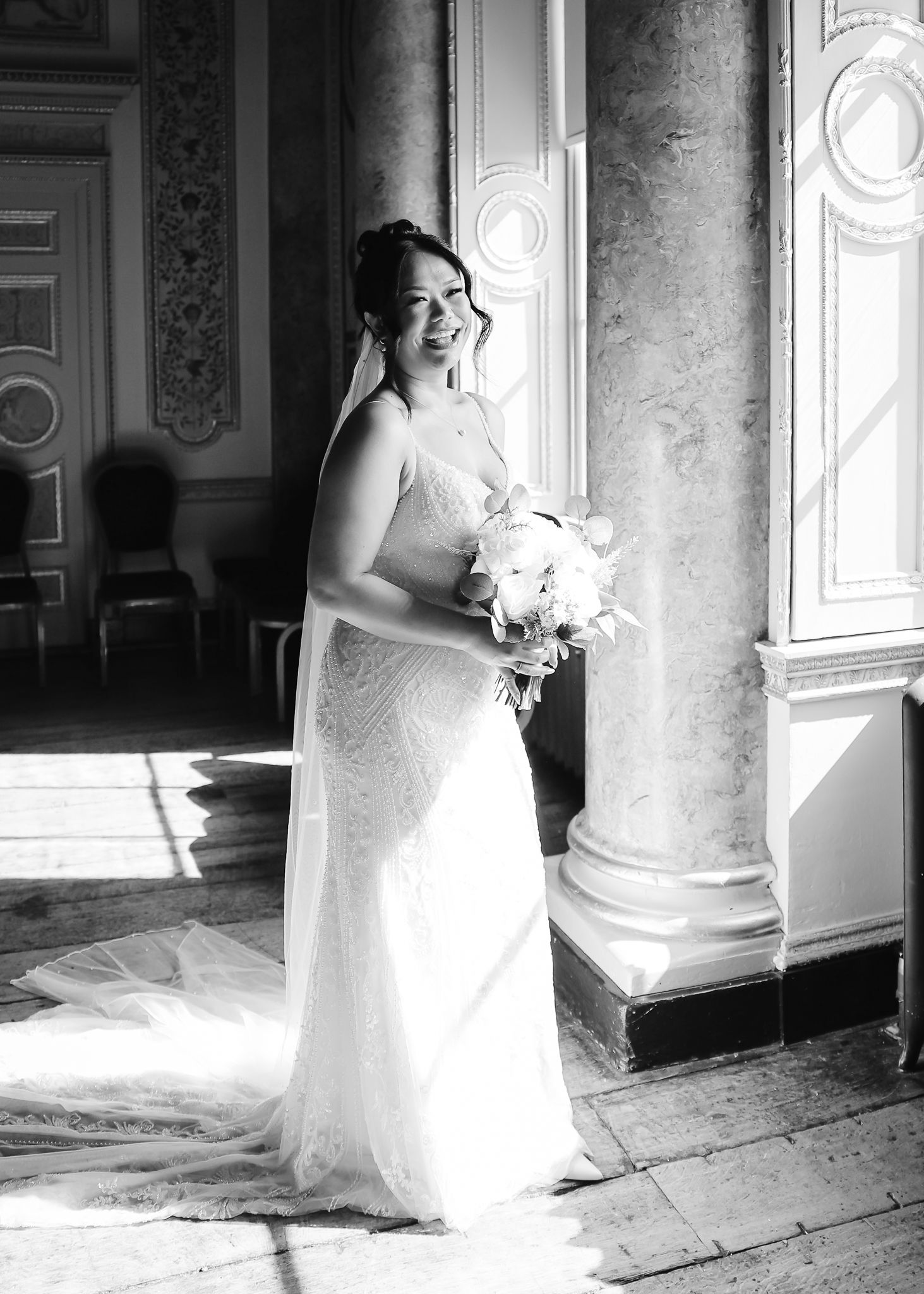 A bride in a white wedding dress poses elegantly by ornate columns and windows in a classic black and white photograph.