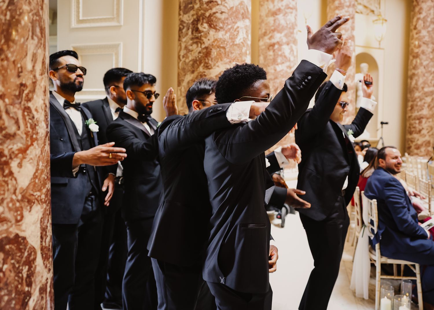 Groomsmen in black suits strike a dramatic pose together in an elegant venue with marble columns and ornate decor.