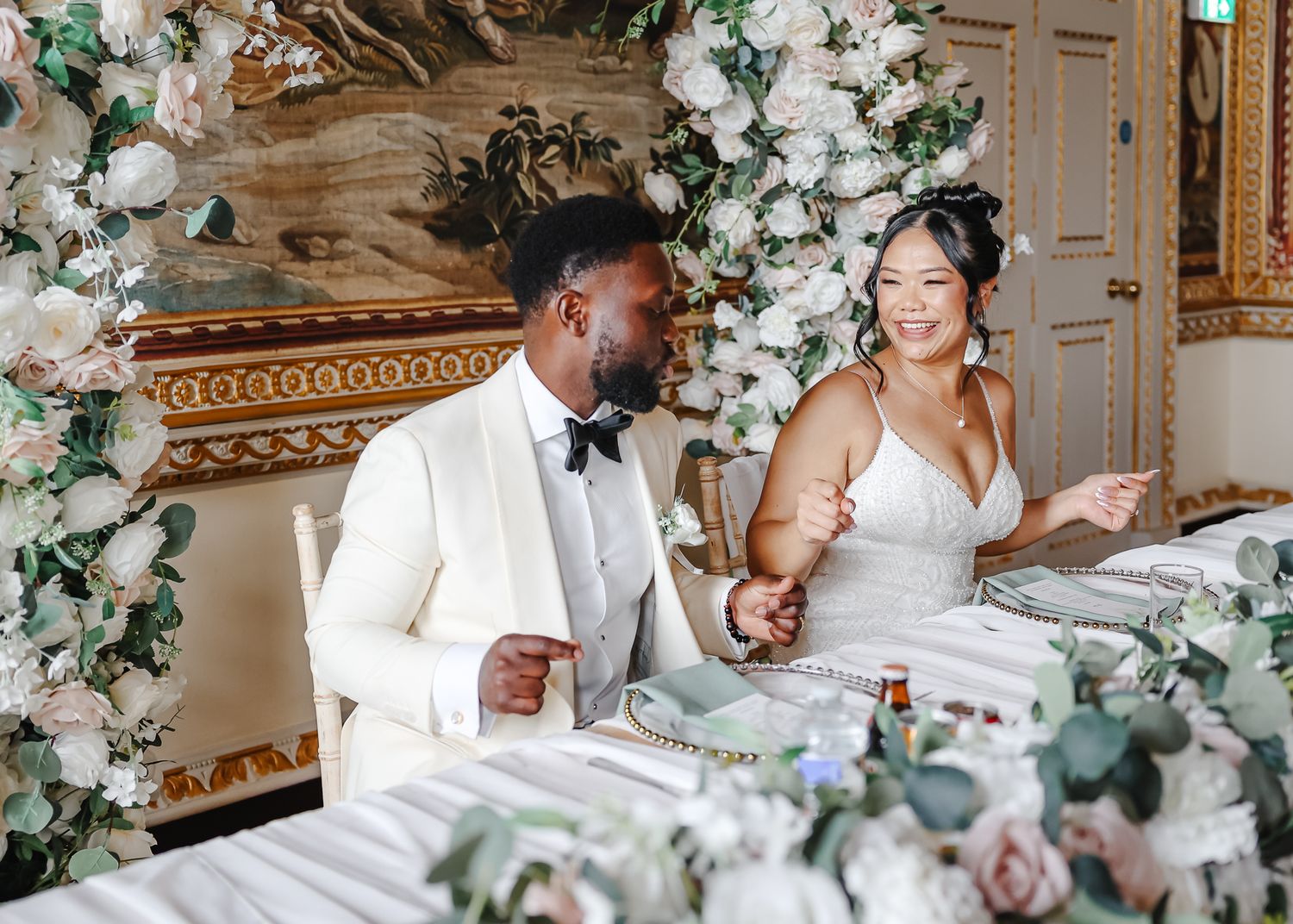 Wedding couple shares a joyful moment while drinking champagne together in an elegant venue decorated with floral garlands.