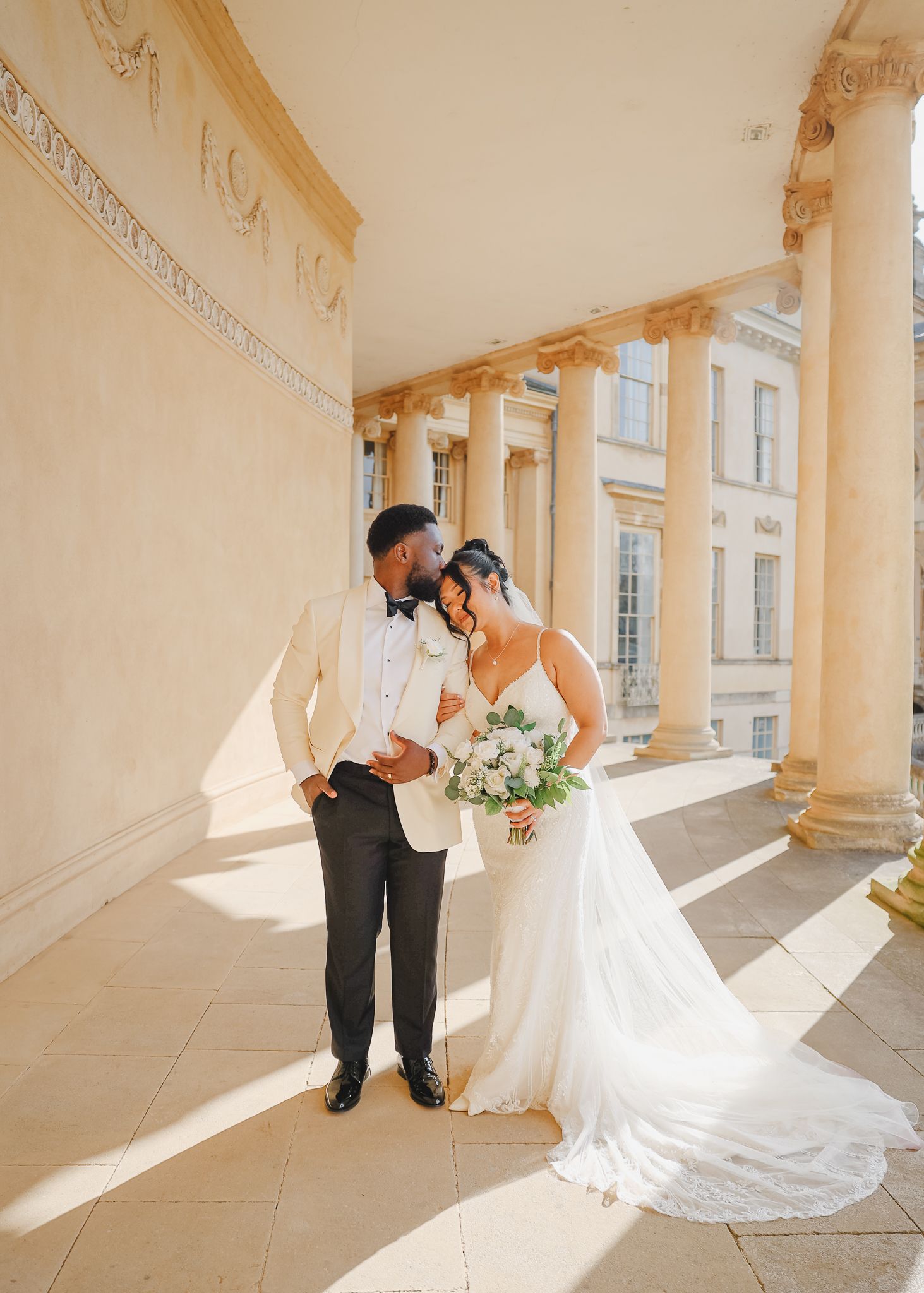 A couple shares an intimate moment in their wedding attire under elegant cream-colored columns with natural lighting.