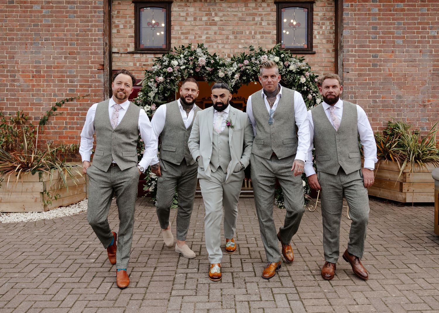 Wedding party in coordinated gray vests and suits pose together on brick patio for group photos.