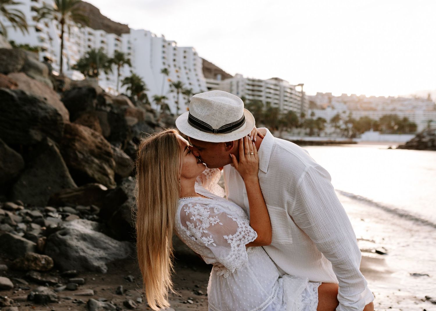 A couple in wedding attire embraces on a scenic rocky beach with ocean waves and palm trees in the background. Gran Canaria