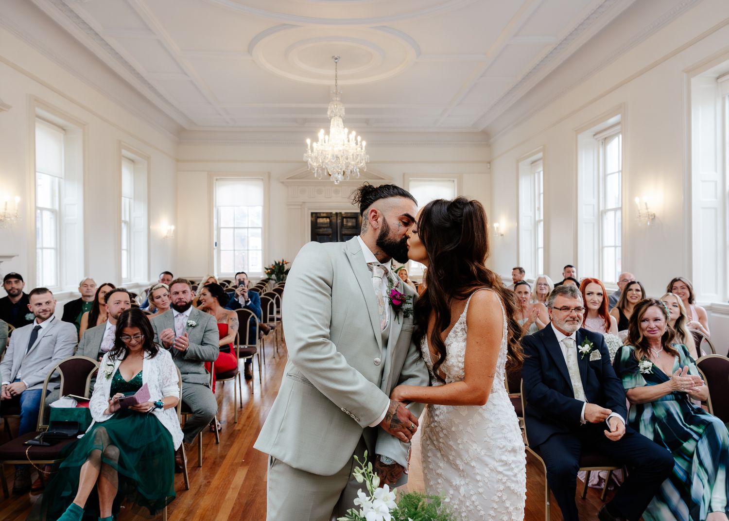 Wedding celebration in a grand white ballroom with guests watching from both sides of the aisle.