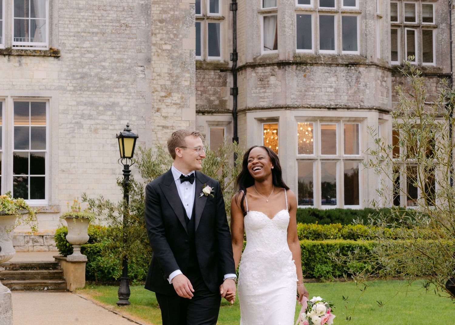 Bride and groom walking hand in hand outside a grand stone manor house on their wedding day.
