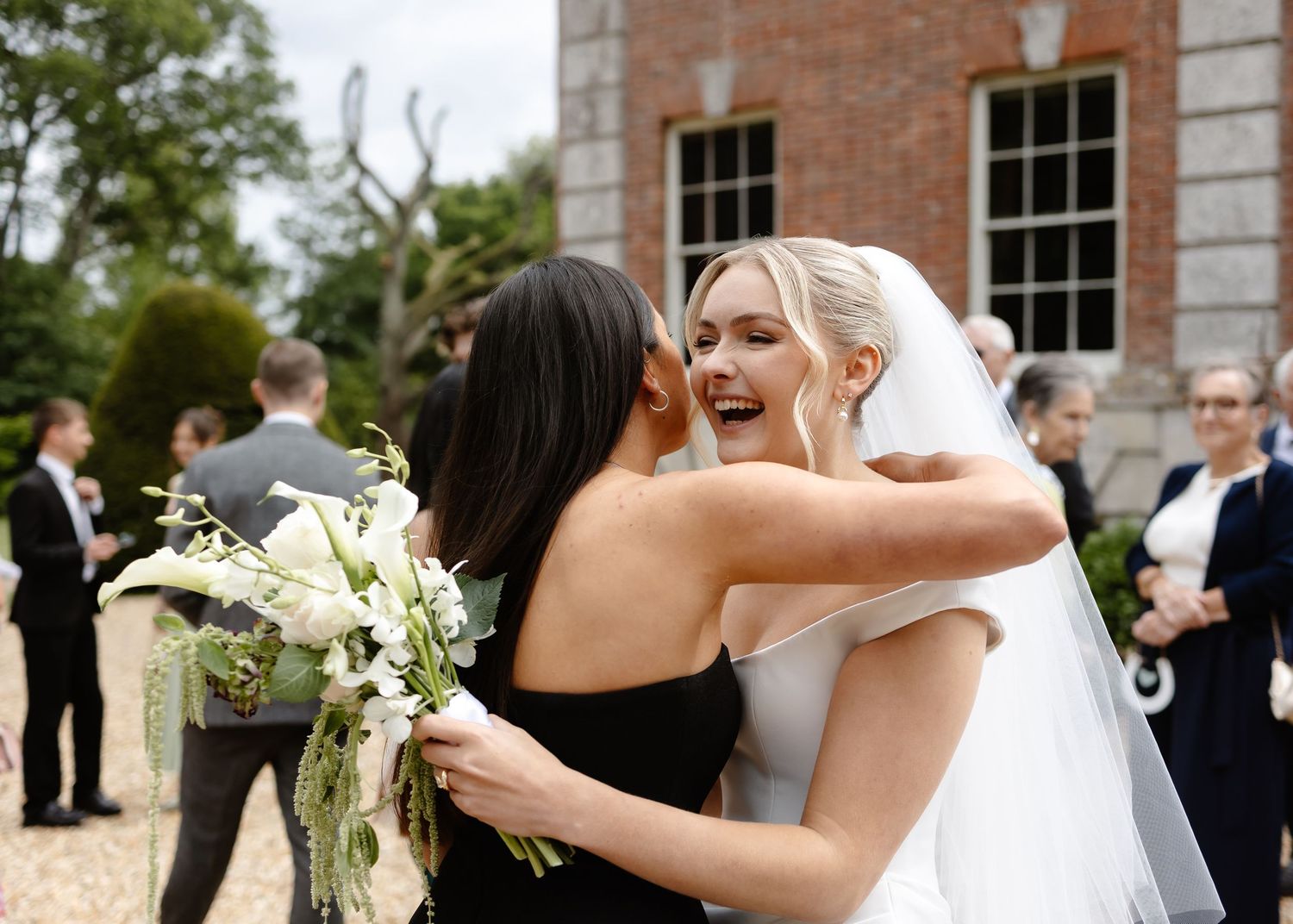 Bride in white veil hugging guest in black dress while holding white floral bouquet outside elegant venue.