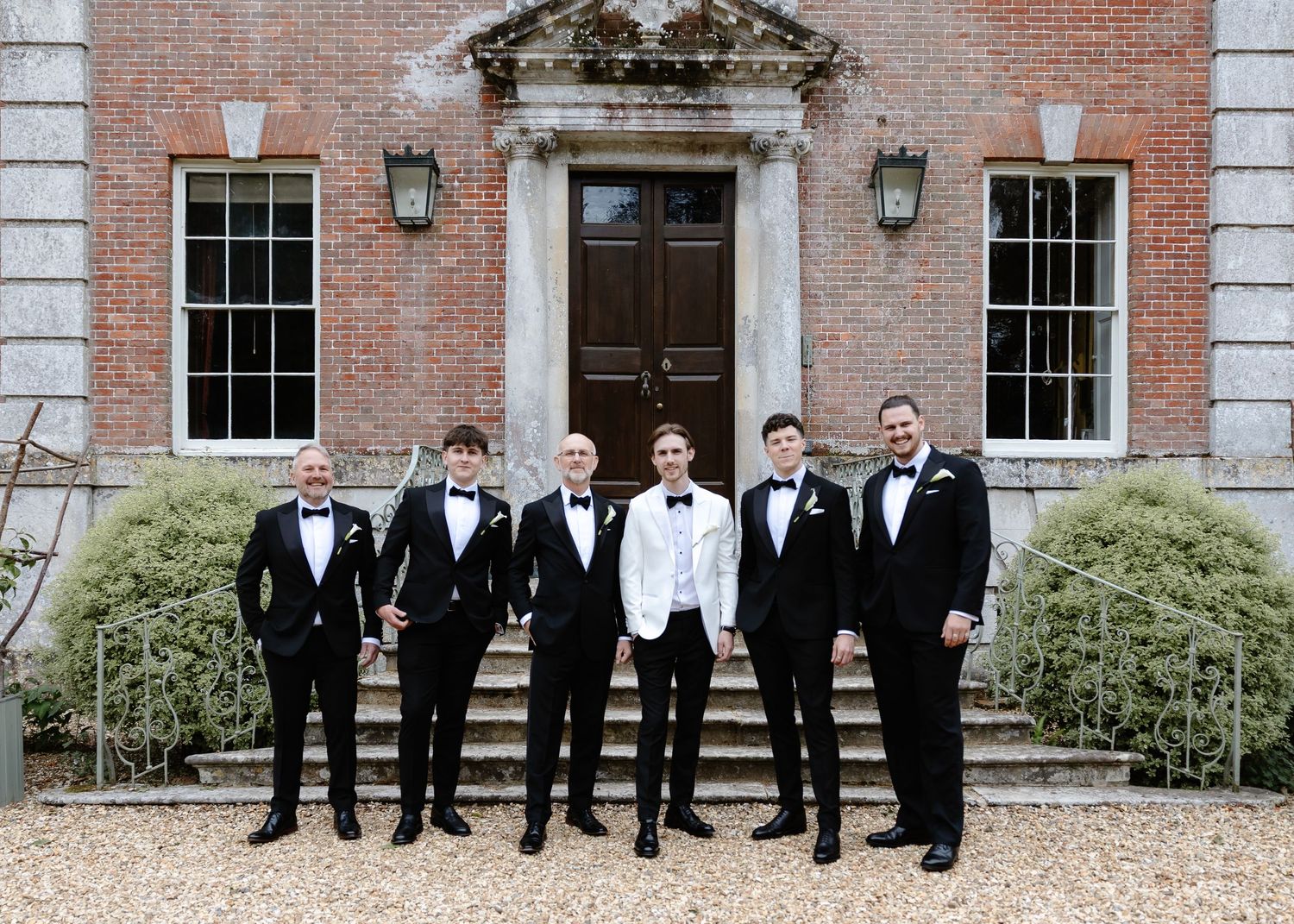Six groomsmen in black tuxedos with one in white jacket pose outside elegant brick manor house for wedding photo.