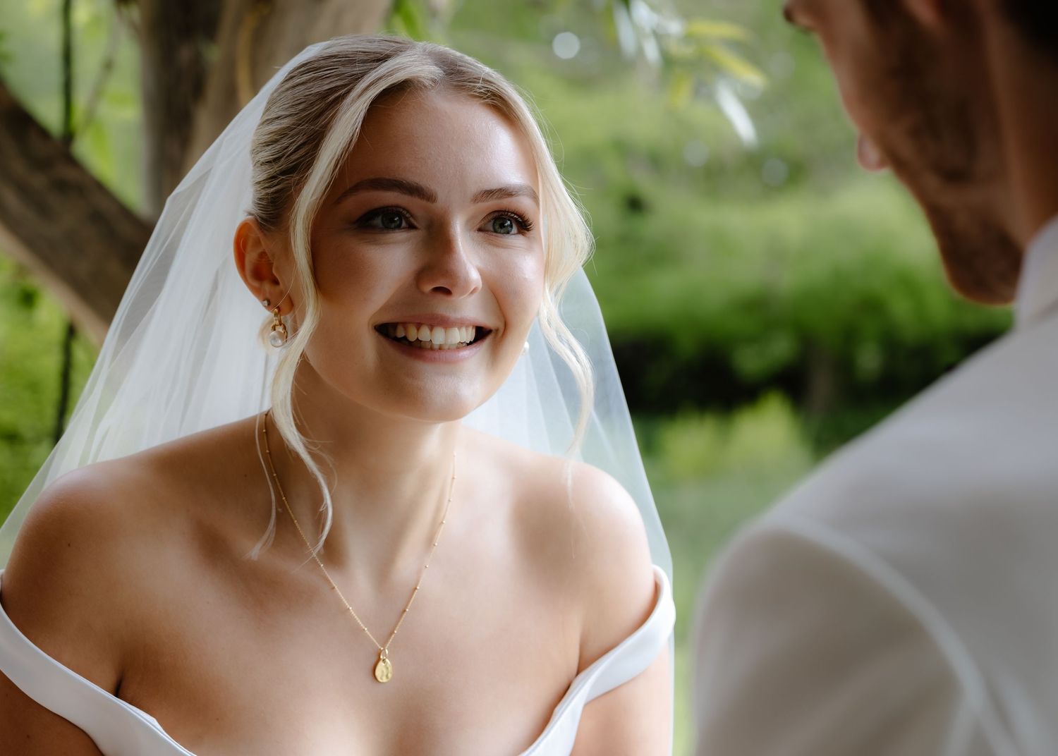 Smiling bride in white veil and off-shoulder gown with gold necklace during outdoor wedding ceremony.