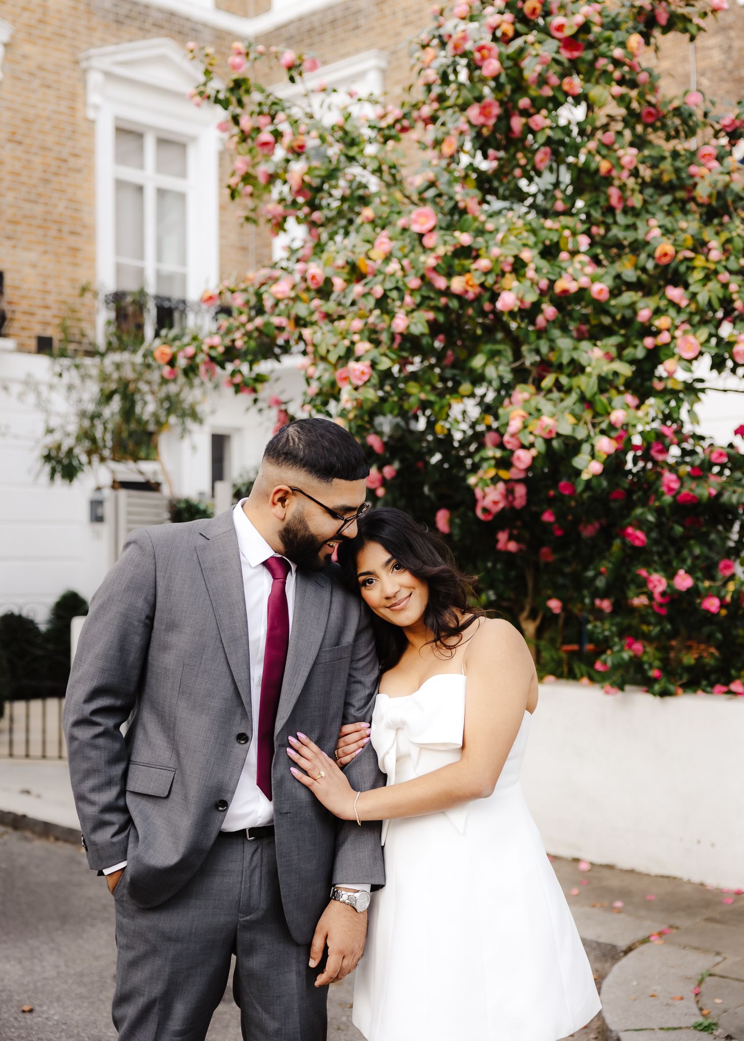 A couple in wedding attire poses in front of a blooming floral tree on a charming street.