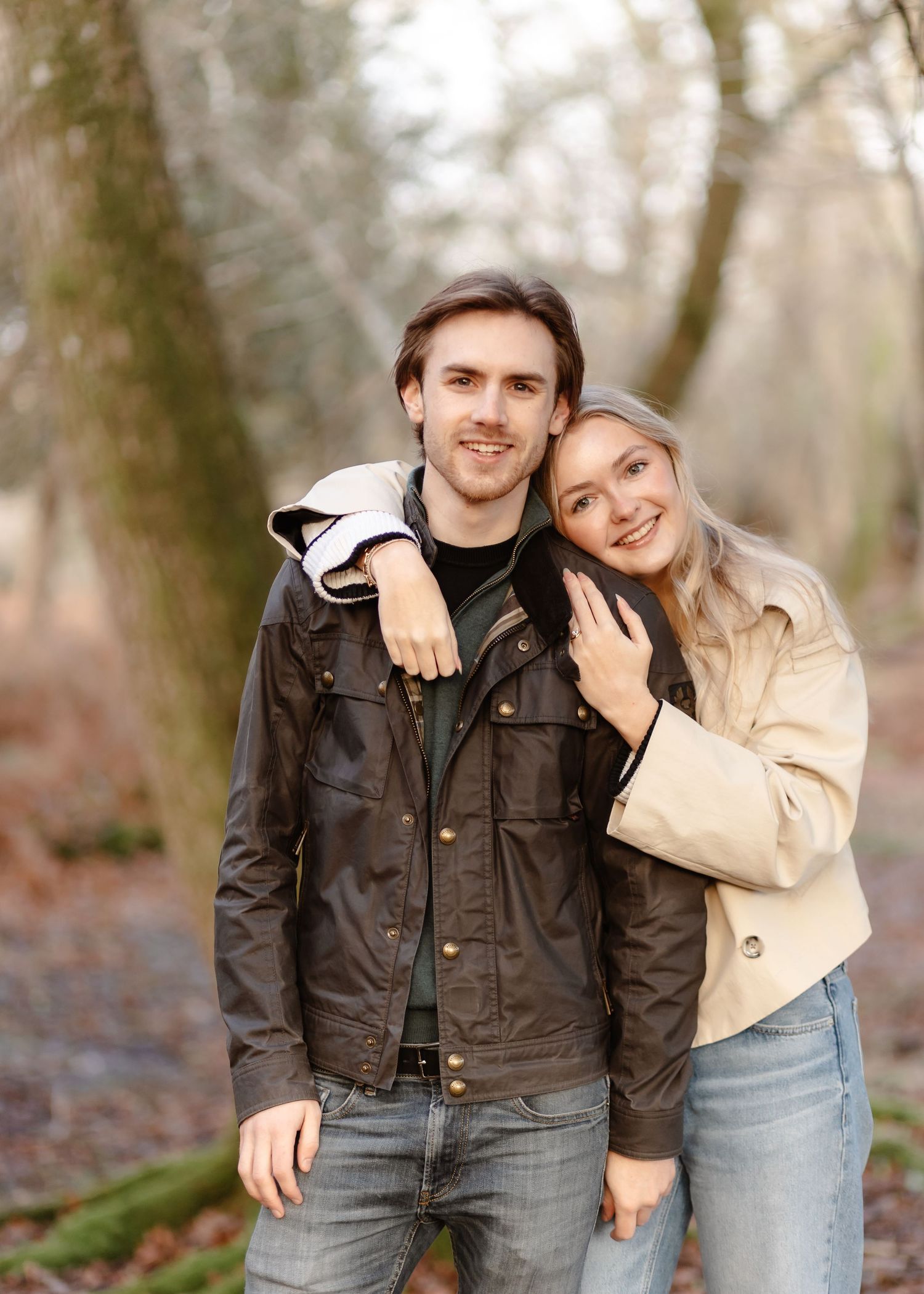 A smiling couple poses together in a woodland setting, the woman embracing the man from behind, both wearing casual jackets.